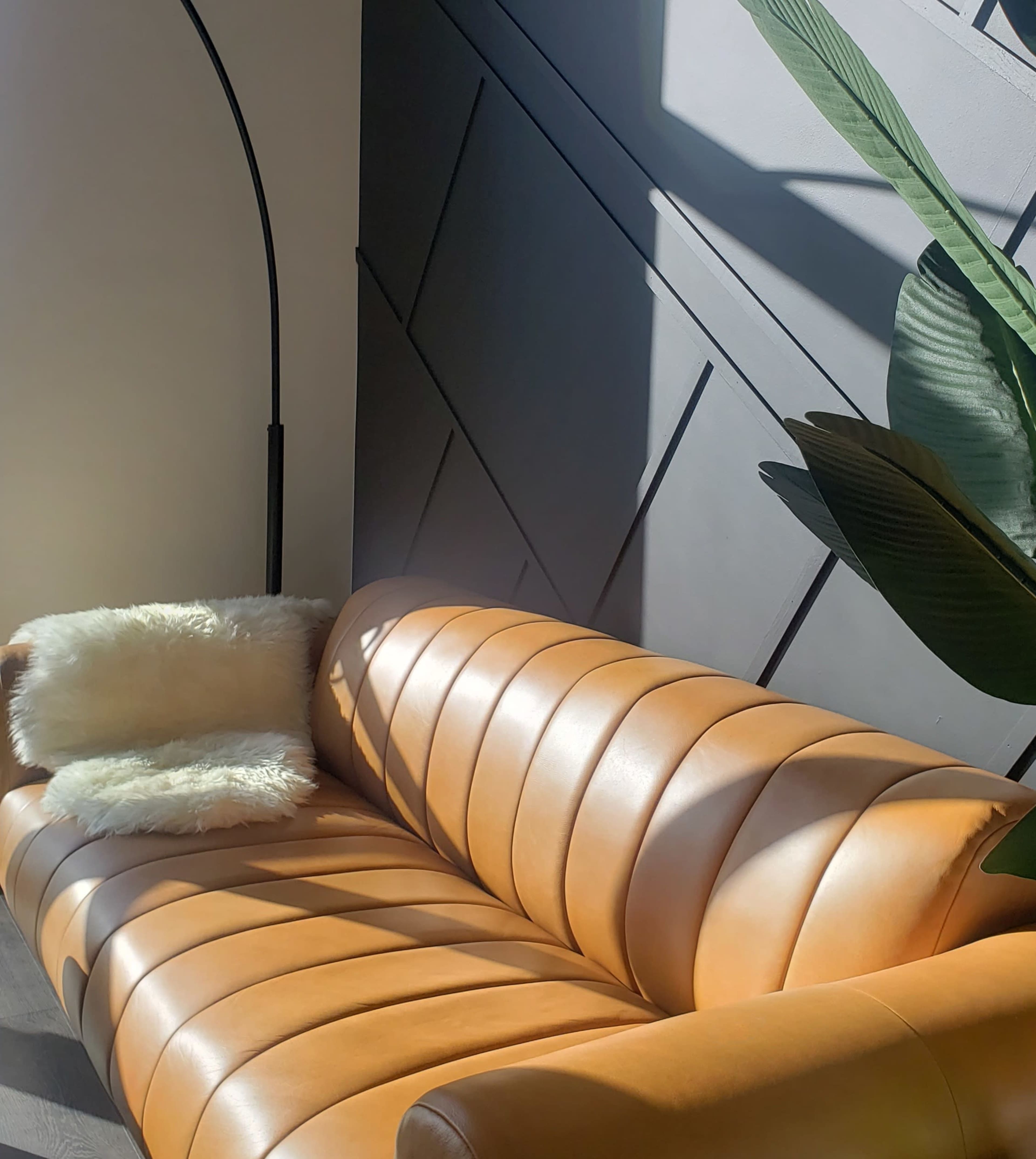A curved orange leather sofa with a white cushion, positioned against a dark gray wall, illuminated by sunlight.