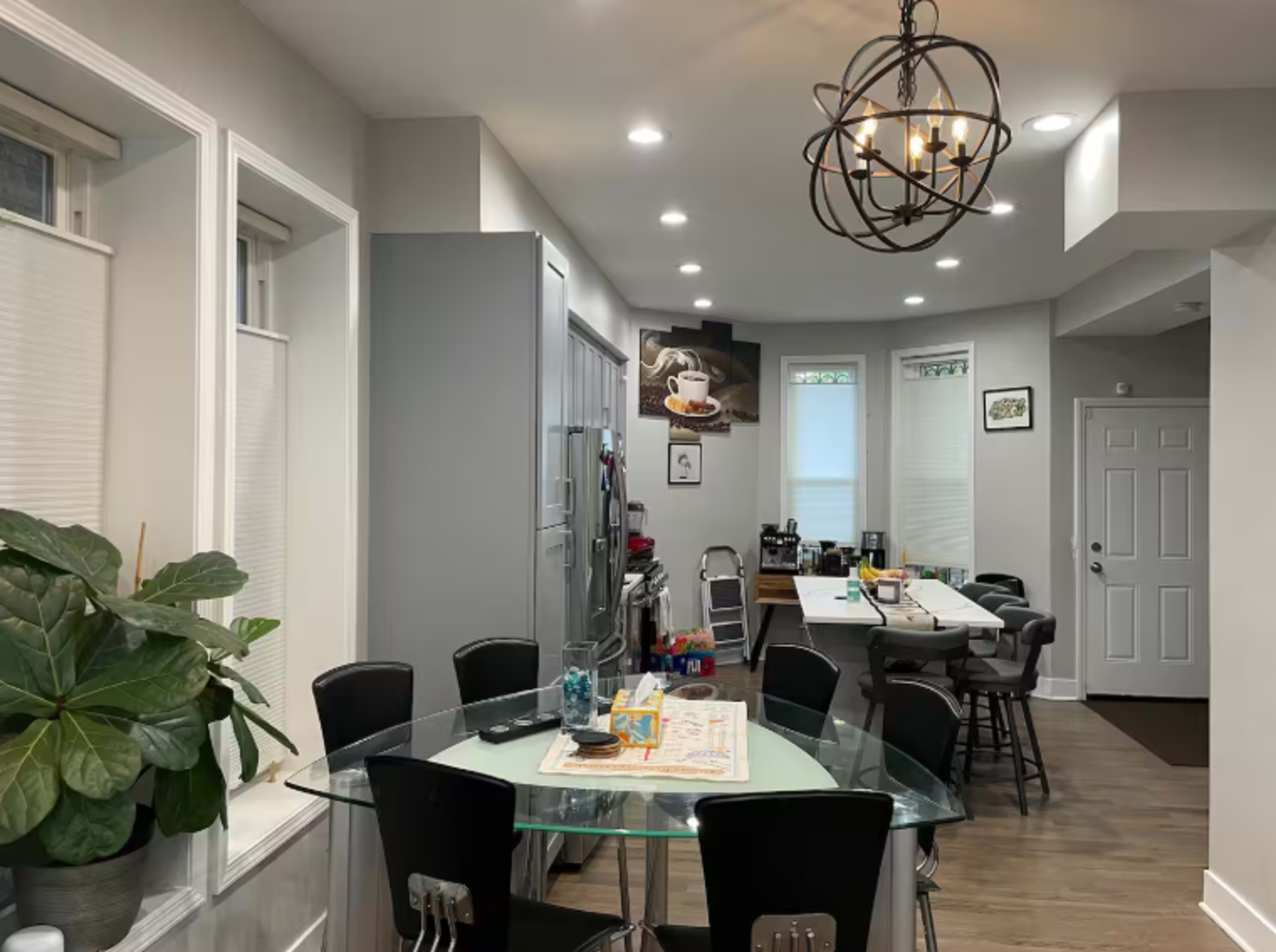 The image shows a modern kitchen and dining area with a glass table, black chairs, and a variety of appliances and décor.