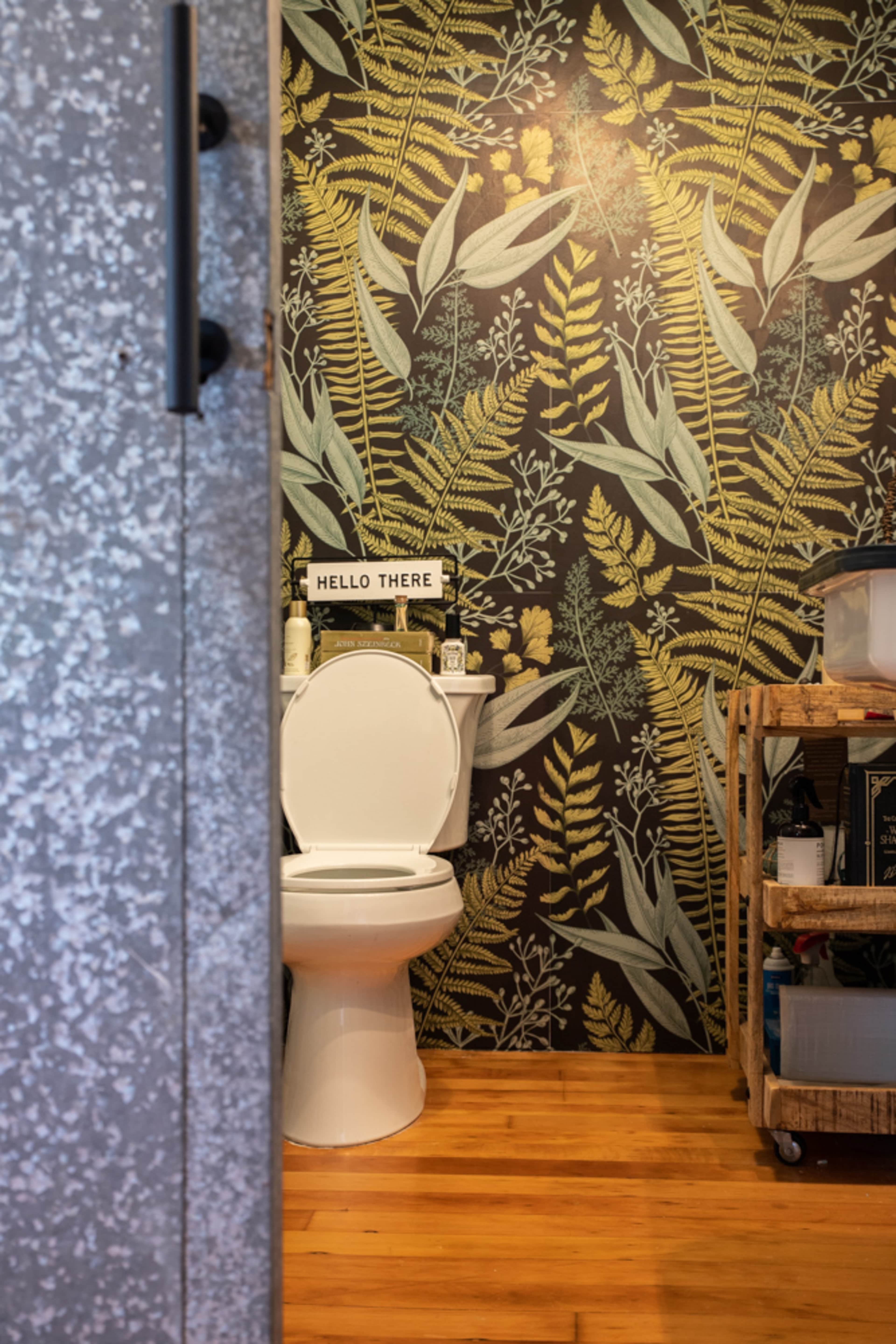A modern bathroom features a white toilet set against a botanical-themed wallpaper and wooden flooring.