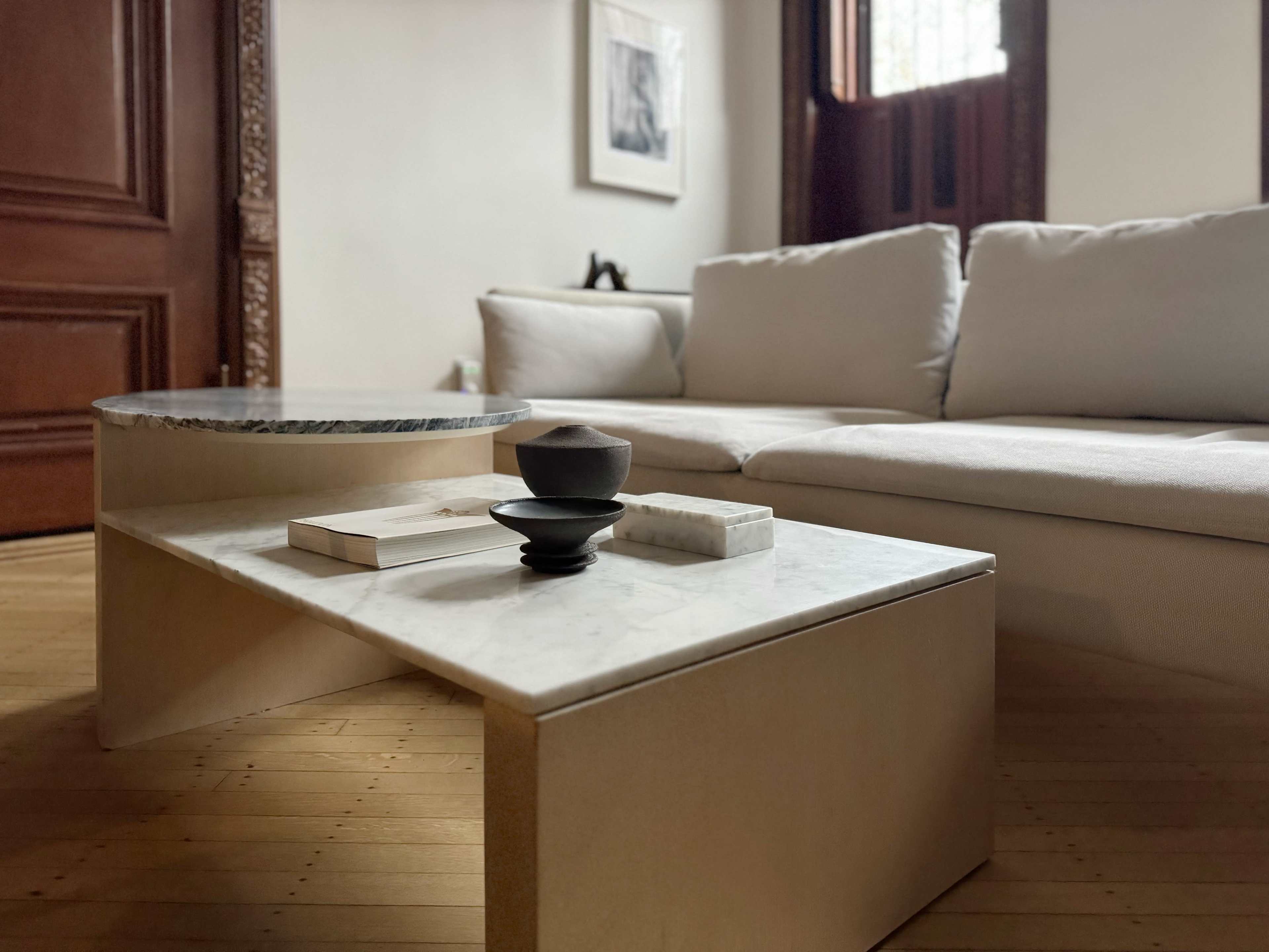 A modern living room features a low, minimalist coffee table with a marble top and a wooden base, surrounded by a light-colored sectional sofa and wooden flooring.
