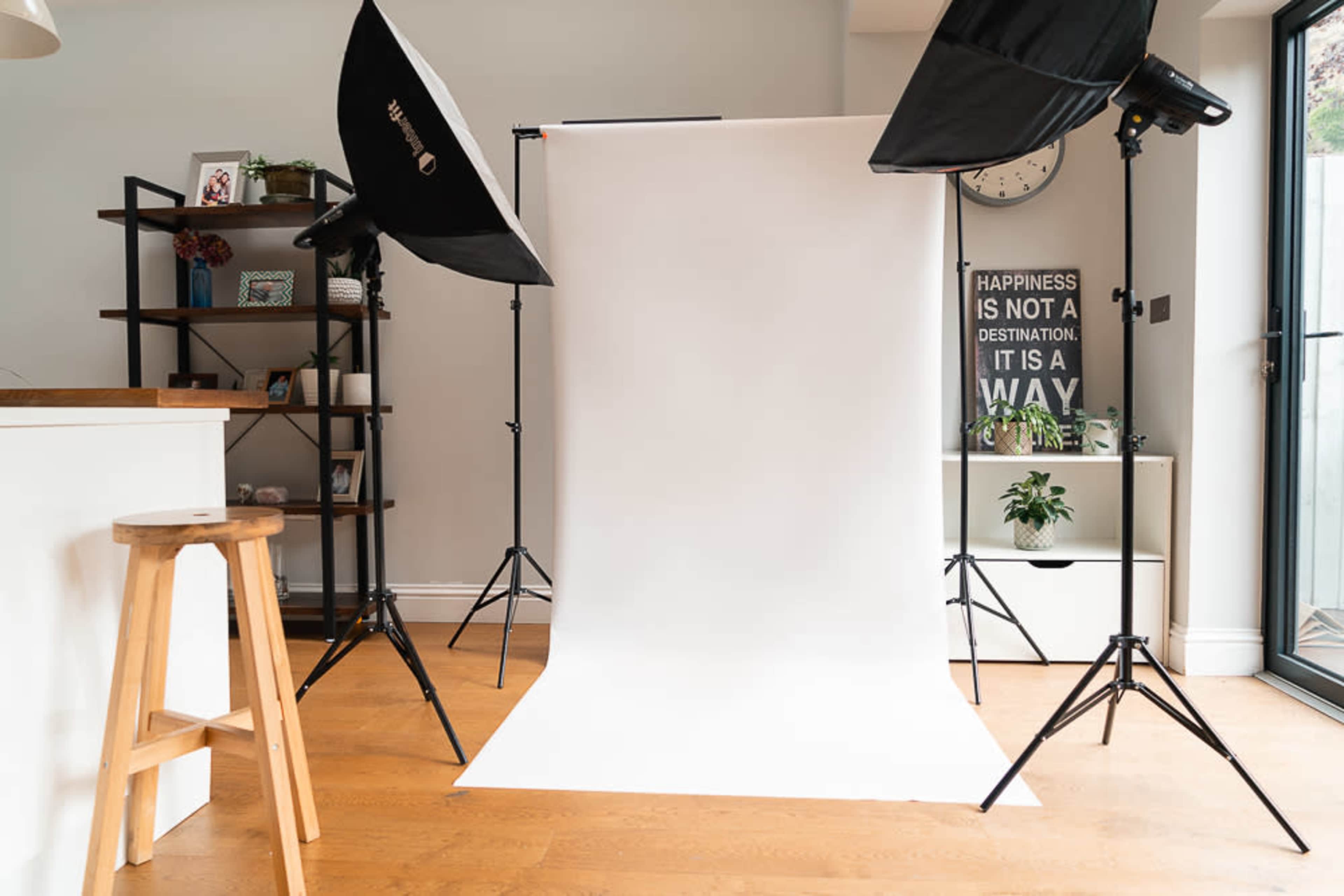 A photography setup features a white backdrop illuminated by two softbox lights in a room with wooden flooring and minimalist decor.