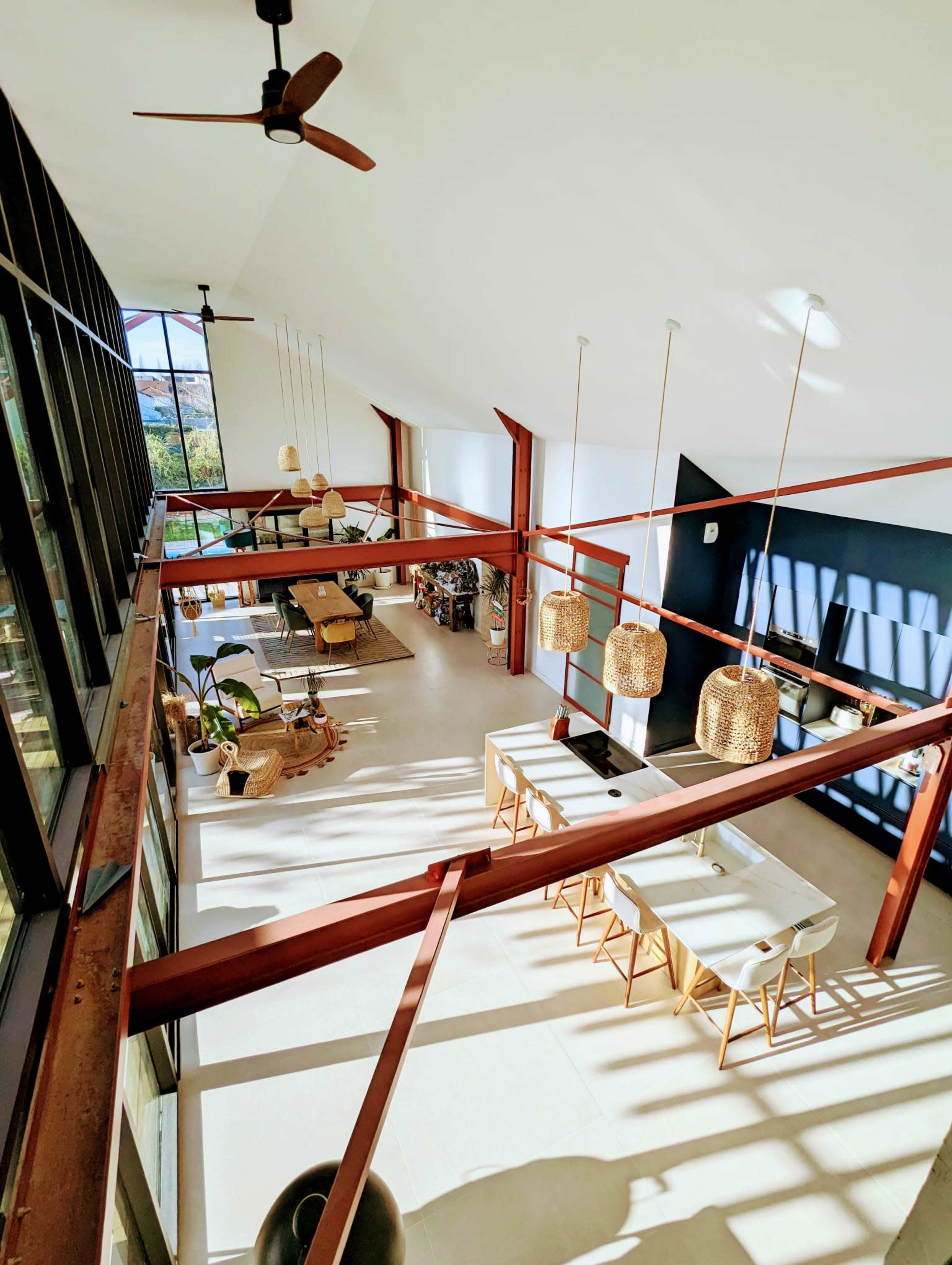 A spacious, modern interior with a high ceiling, large windows, and a dining area featuring a long table and hanging light fixtures.