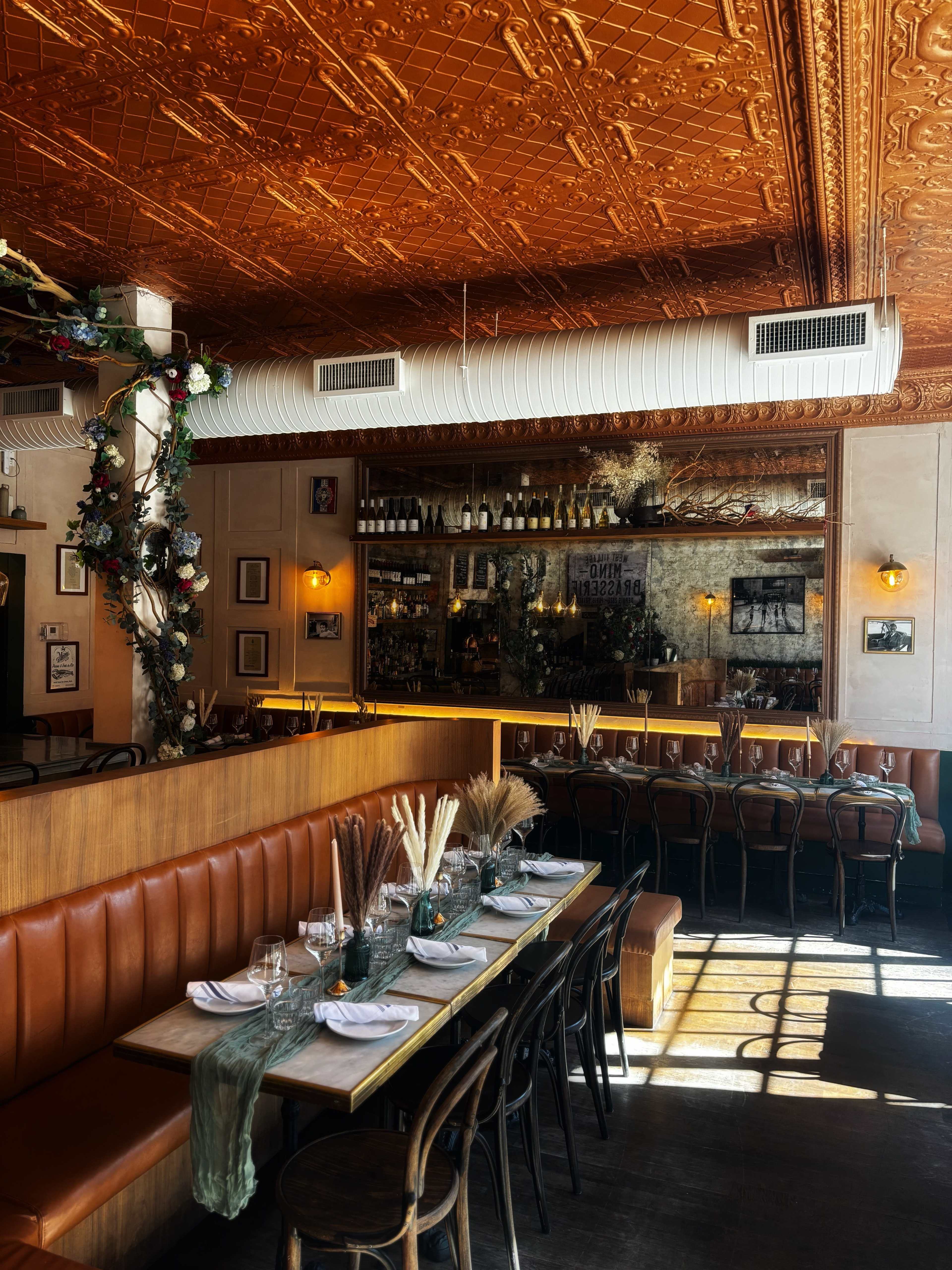 The image shows an inviting restaurant interior with a richly detailed ceiling, brown leather booths, and a long table set for dining, illuminated by natural light.