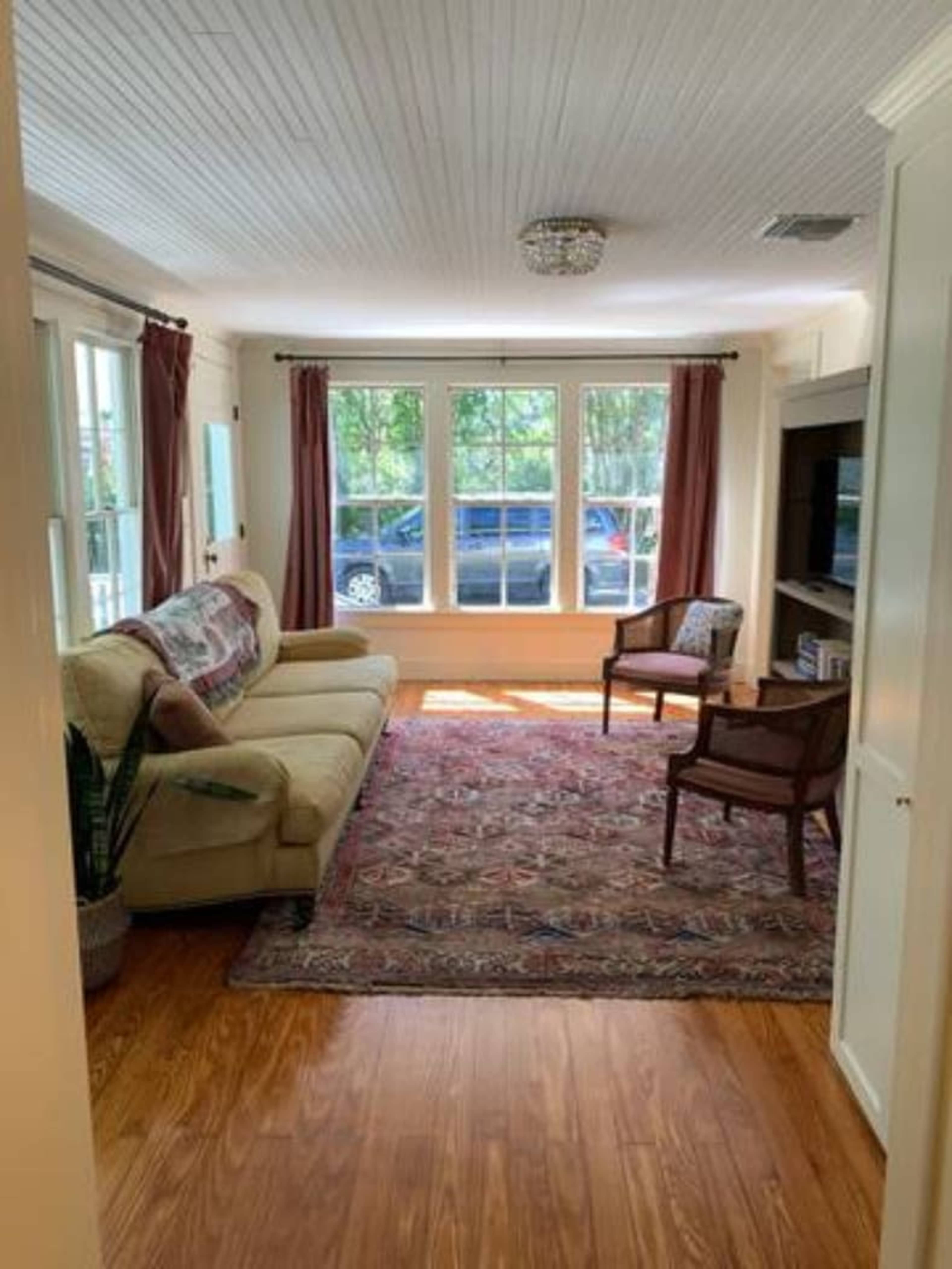A room with a beige sofa, a patterned rug, and two chairs, illuminated by natural light from large windows.