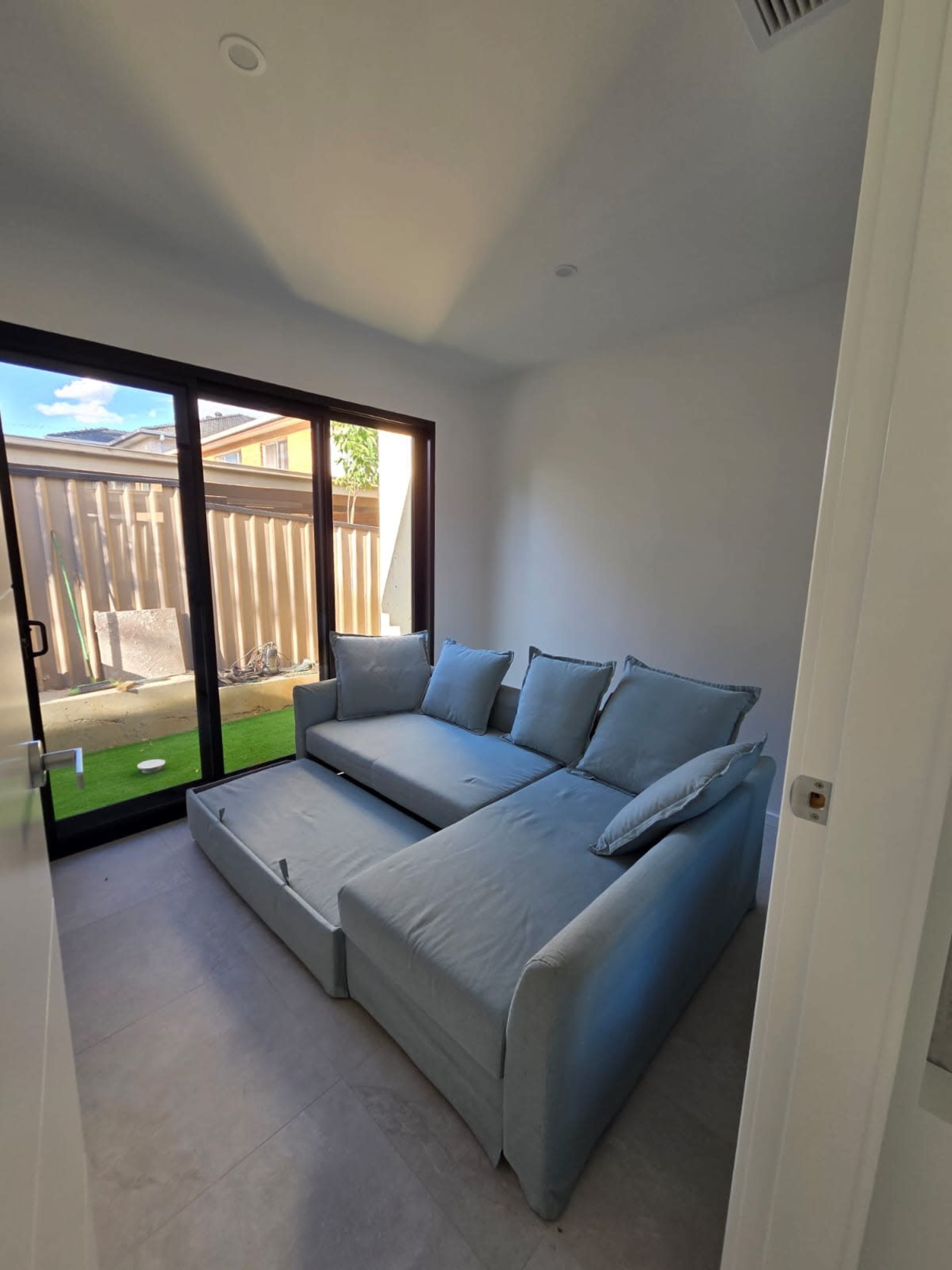 The image shows a simple, modern room with a light blue sectional sofa and large glass doors leading to an outdoor area.