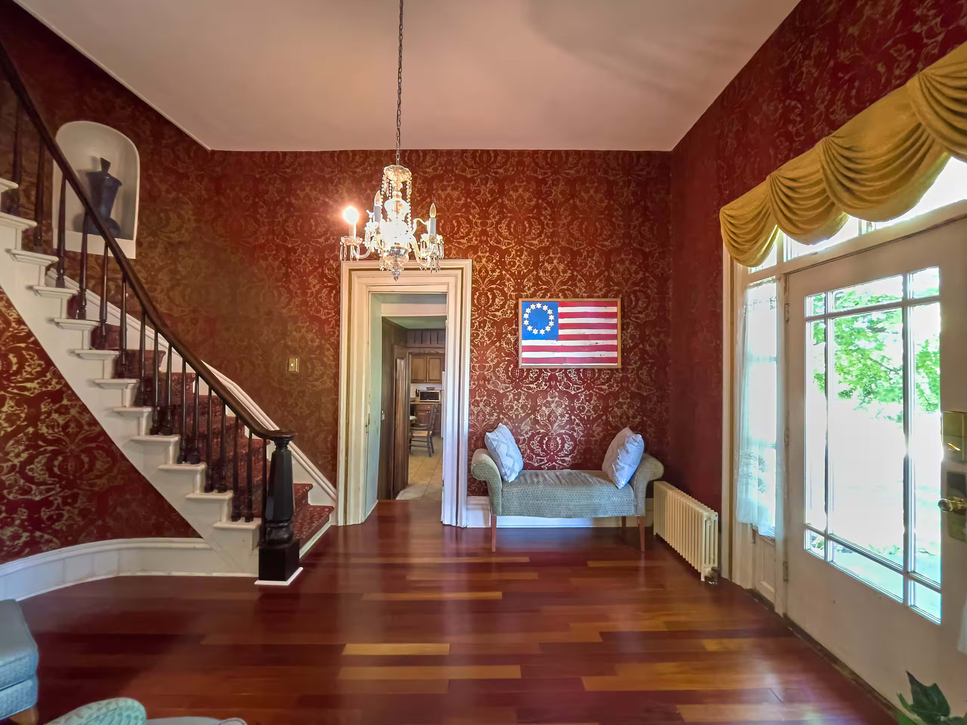 A grand foyer features a staircase with dark wood railings, a chandelier, and a decorative red wallpaper, illuminated by natural light from a large window.