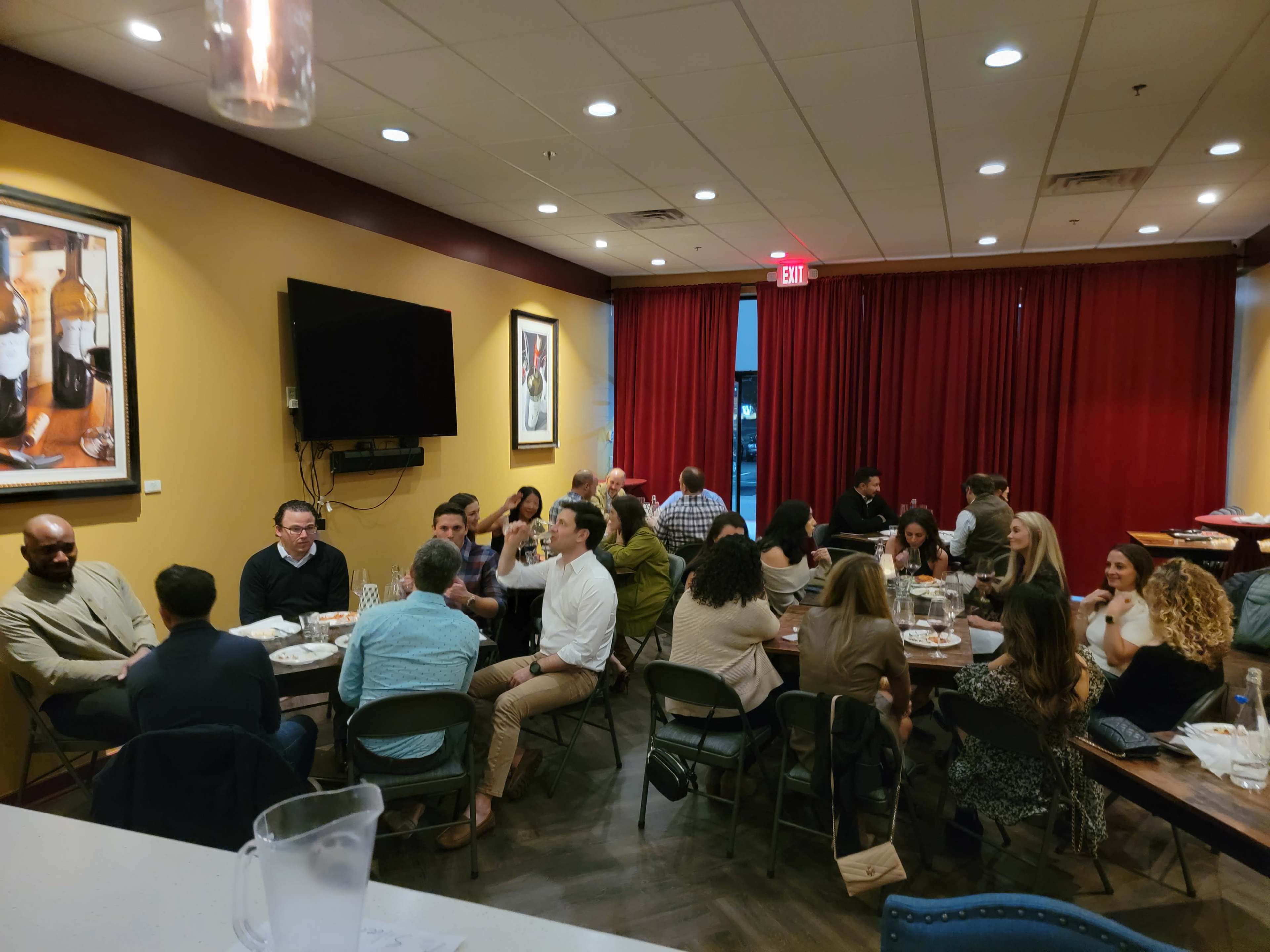 A group of people is gathered around tables in a restaurant, engaged in conversation, with red curtains and a large wall-mounted television in the background.