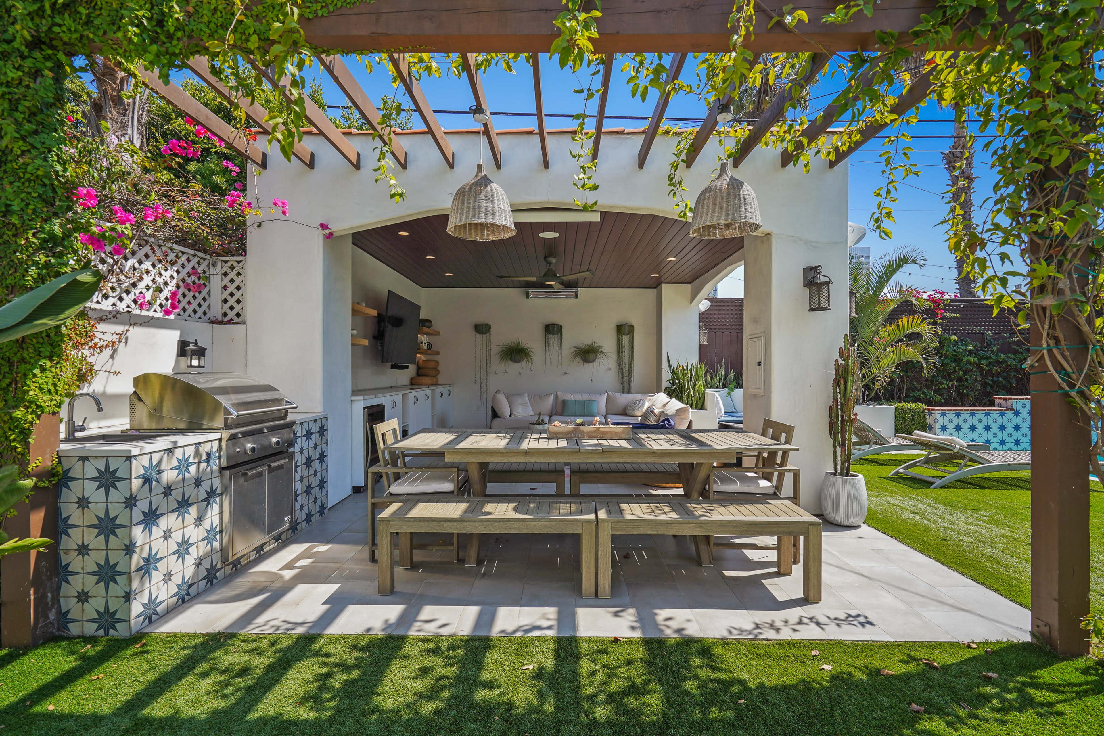 The image shows an outdoor kitchen and dining area with a large wooden table, stainless steel grill, and decorative tile accents under a pergola.