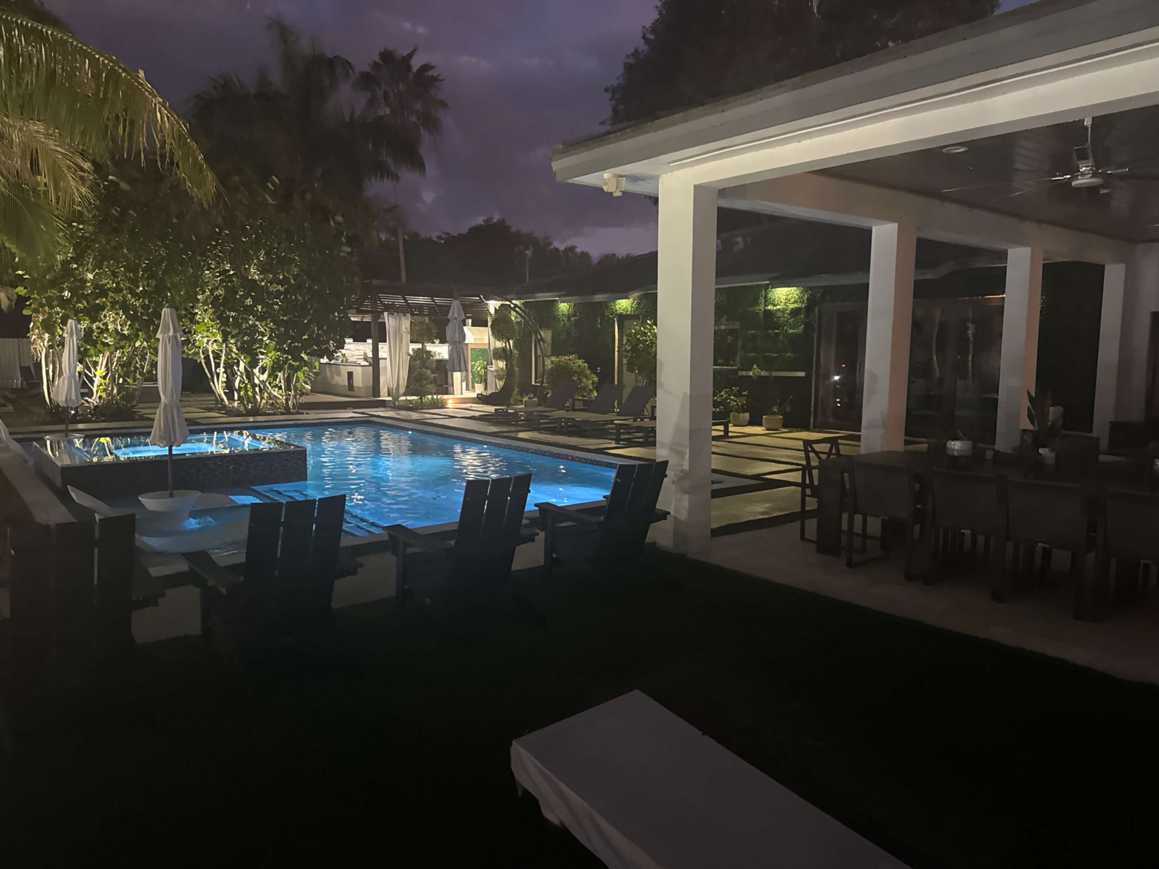 A nighttime view of a pool area surrounded by lounge chairs, palm trees, and a house with illuminated features.