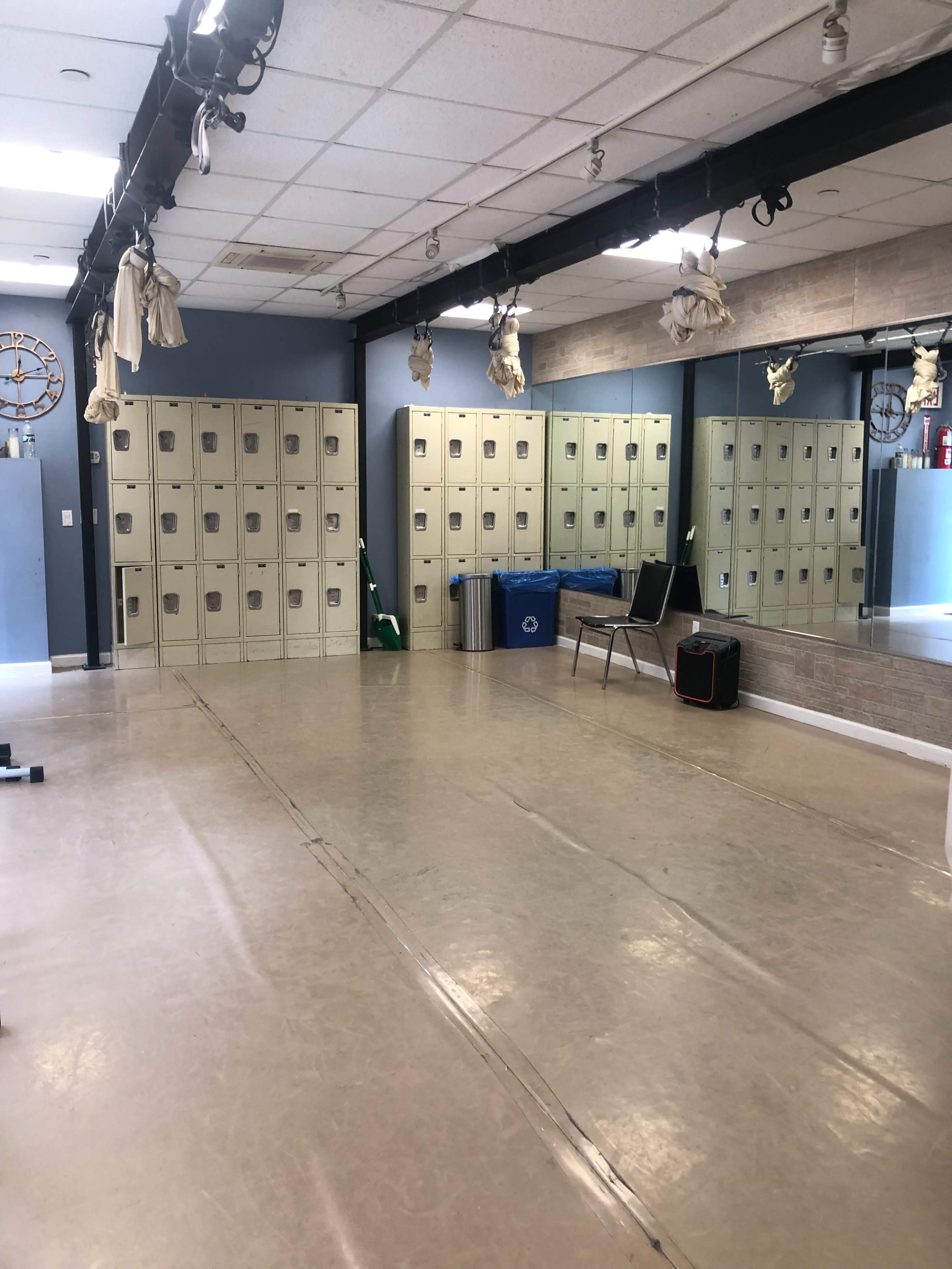 The image shows a spacious room with rows of beige lockers on one side, a mirror reflecting the opposite wall, and minimal furniture, including a chair and a small bag on the floor.