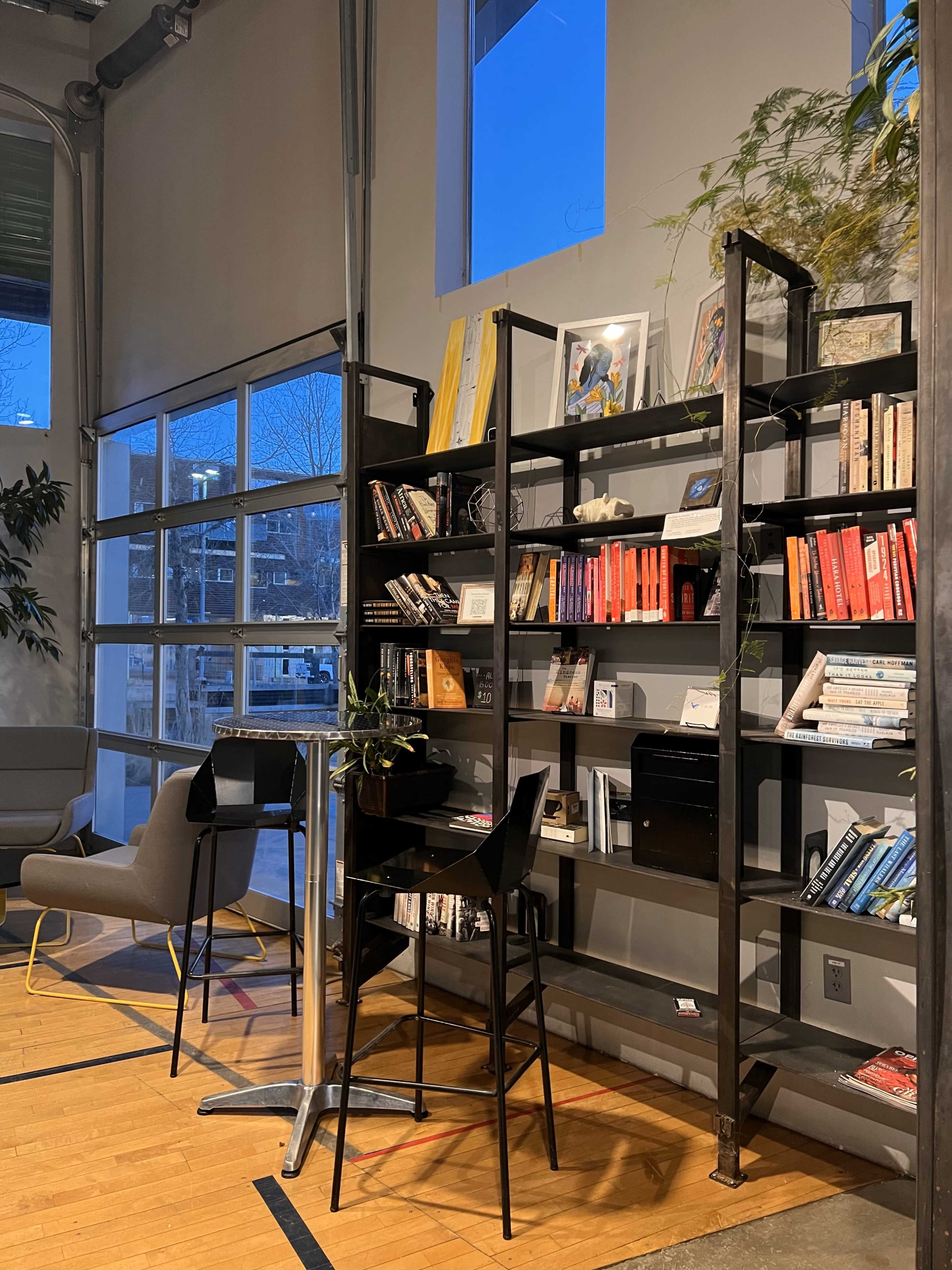 A modern indoor space with a metal bookshelf filled with books, a small table, and chairs, illuminated by soft evening light coming through large windows.