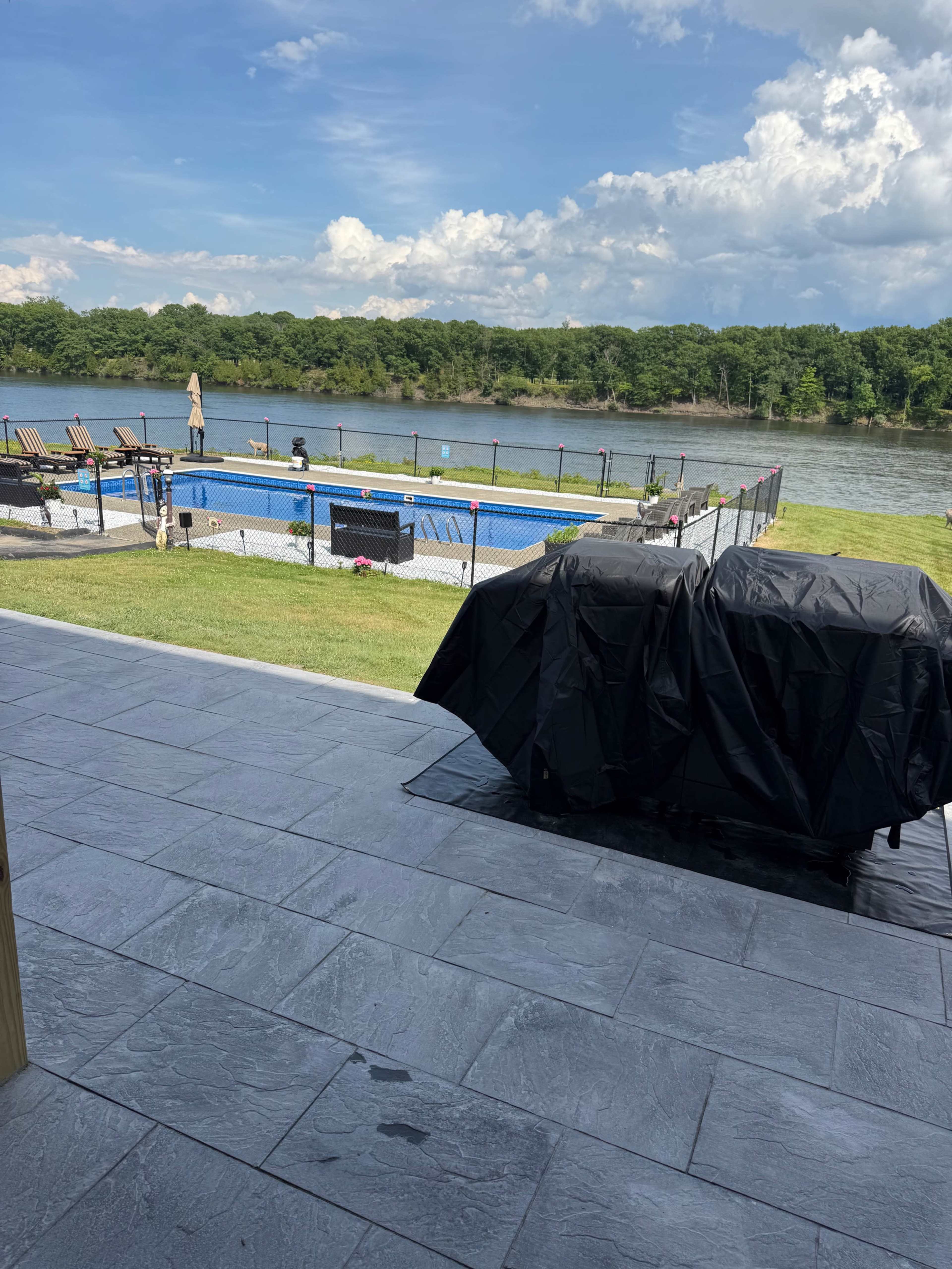 The image shows a view of a backyard with a swimming pool, patio, and a river beyond, set against a backdrop of trees and partly cloudy skies.