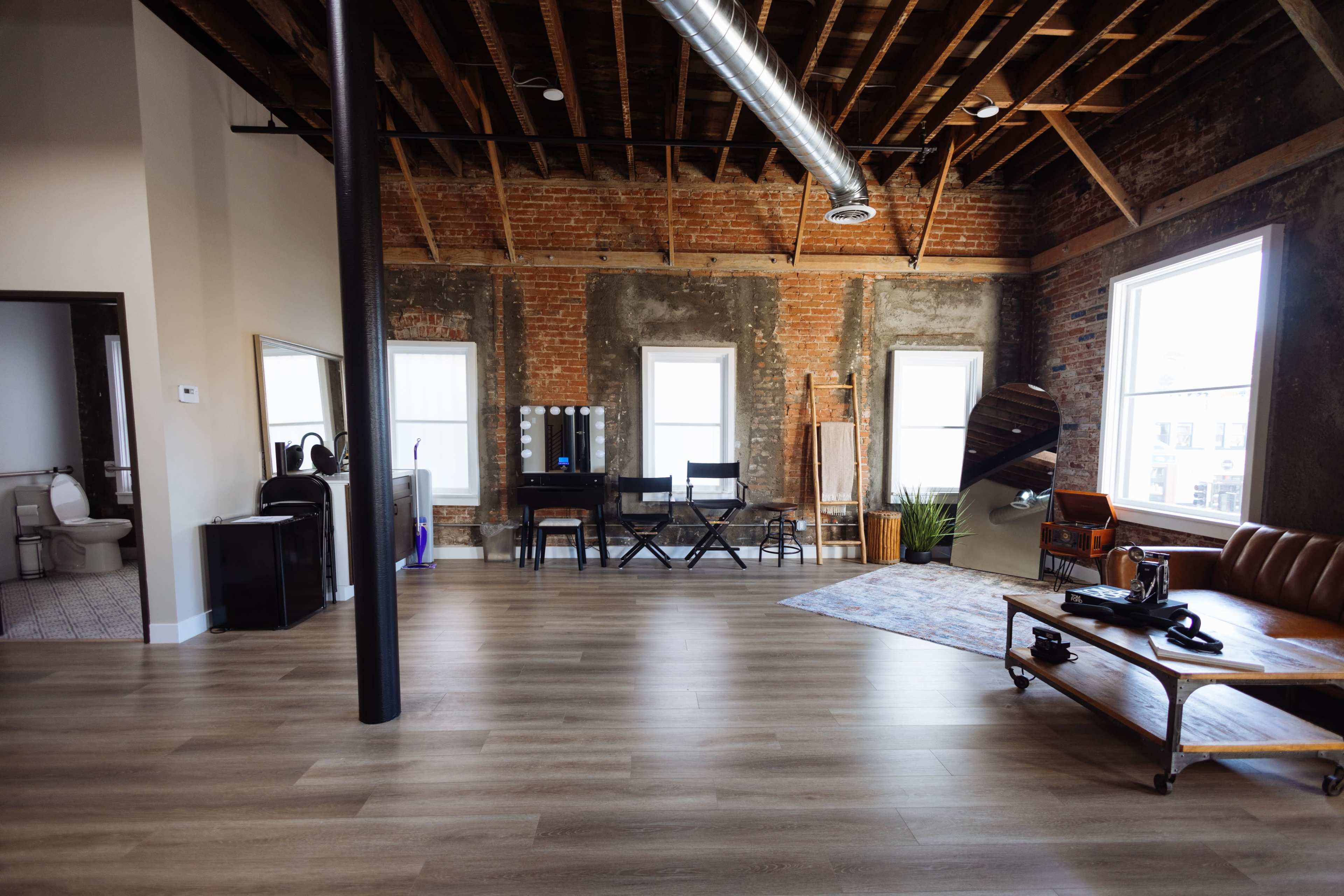 A spacious loft-style interior with exposed brick walls, wooden beams, a wooden floor, and a mix of furniture including a brown leather sofa, dining chairs, and a small table.