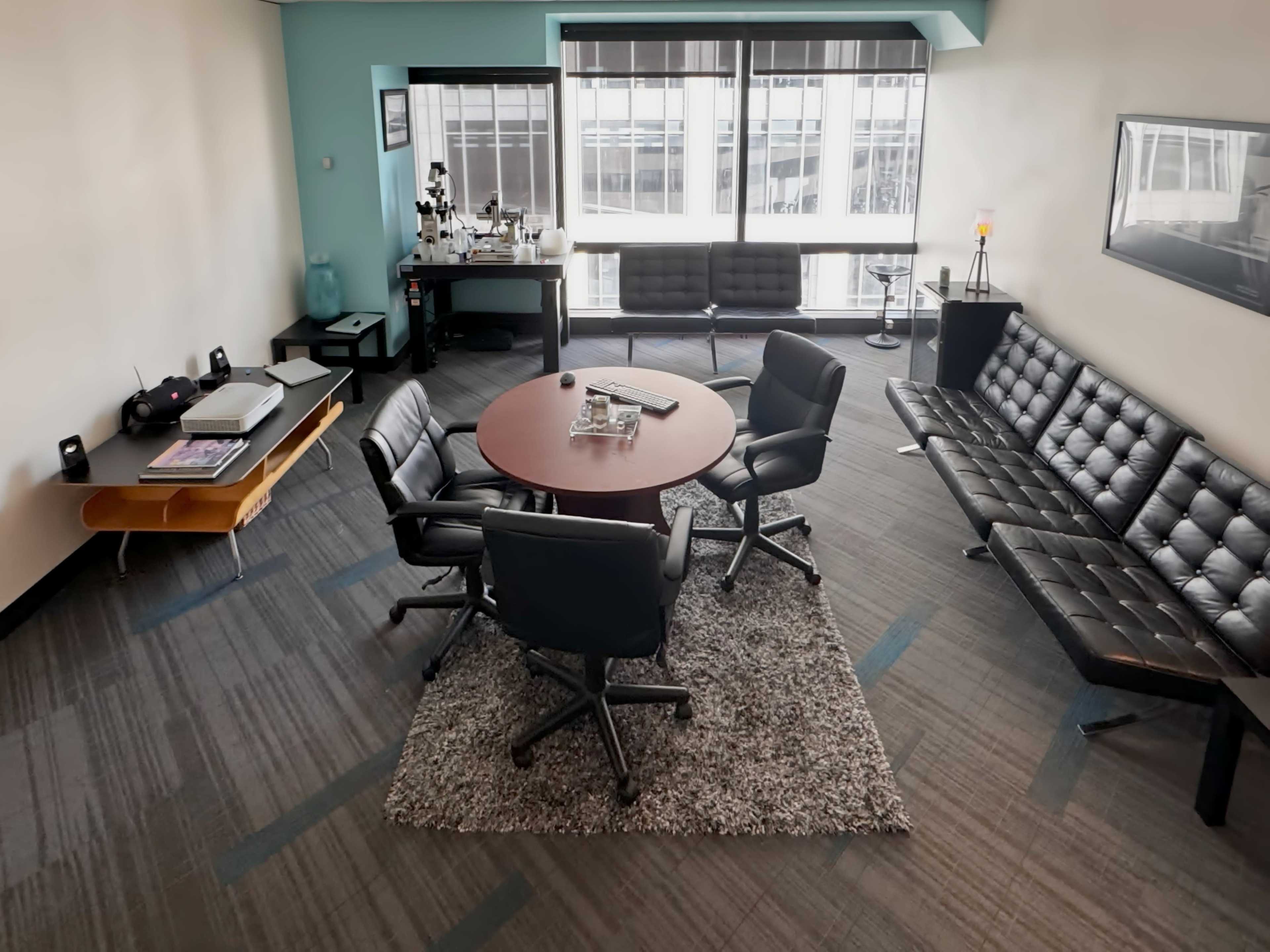 A modern office space featuring a round meeting table surrounded by black ergonomic chairs, a gray area rug, and seating along the walls.