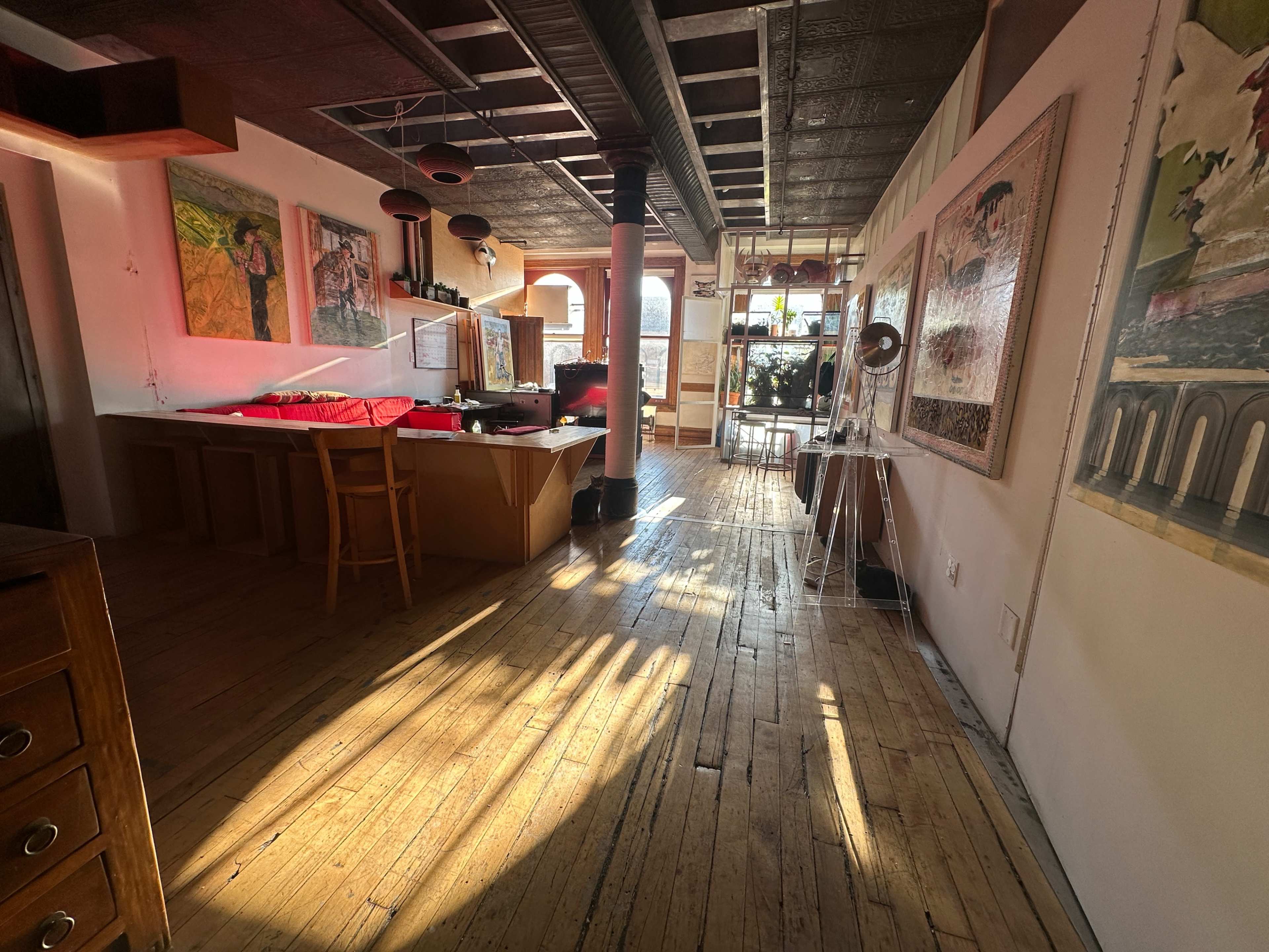 The image shows a spacious interior with wooden flooring, large windows allowing natural light, and several artworks displayed on the walls.