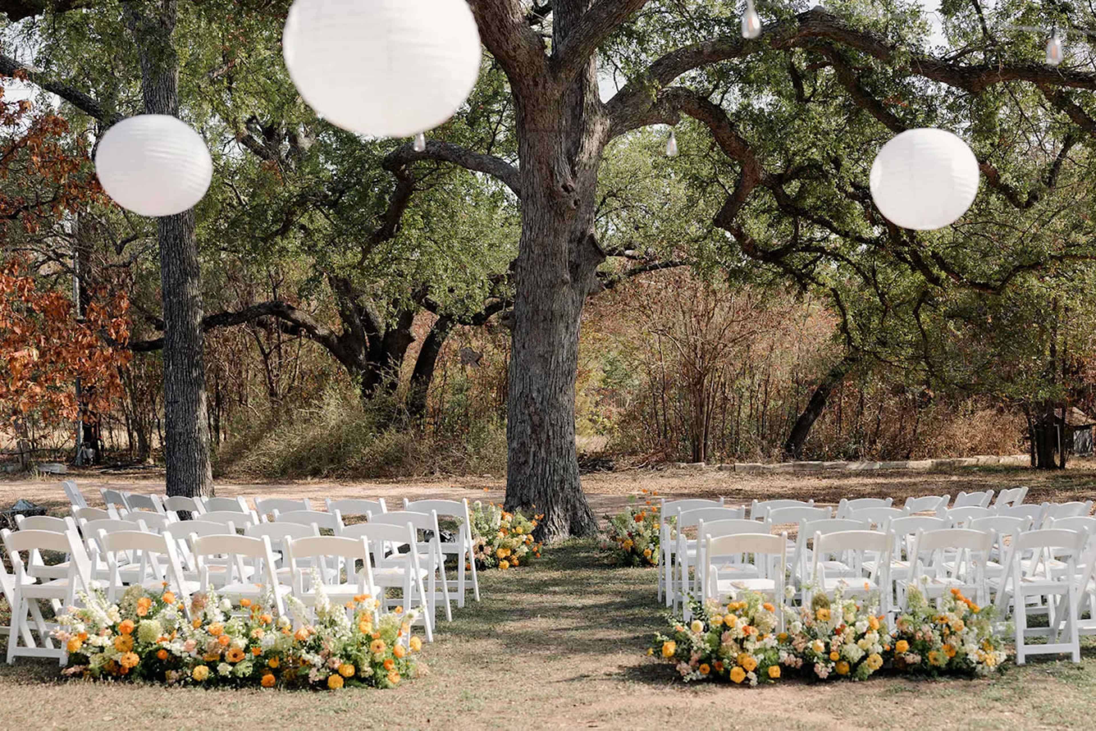 The scene features a wedding ceremony setup with white chairs arranged in rows, surrounded by floral arrangements and hanging lanterns under a large tree.