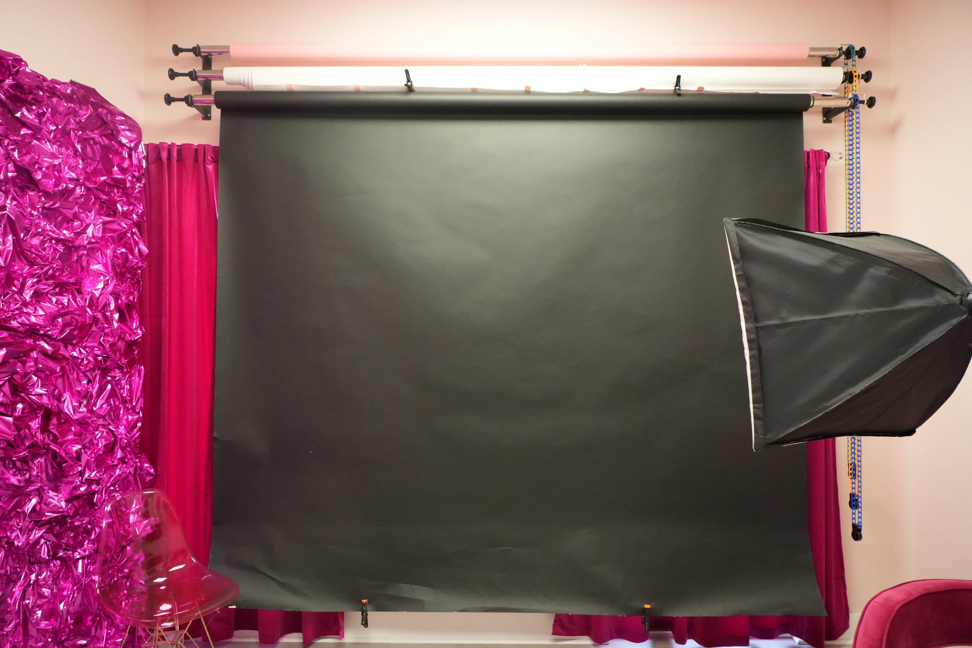 The image shows a photography studio with a black backdrop, a softbox light on the right, and pink curtains and decorations in the surrounding area.