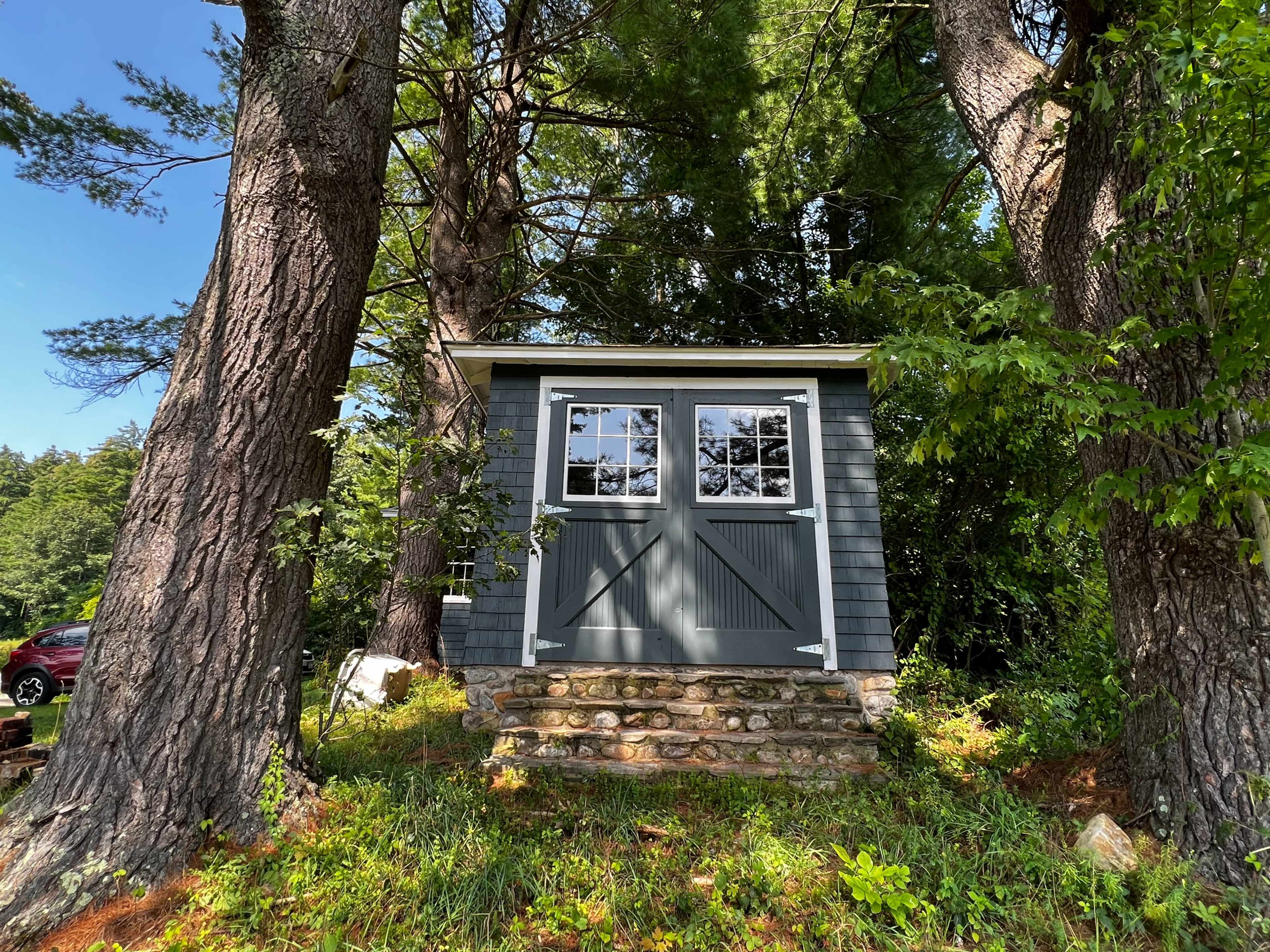 A small, gray shed with double doors is situated on a stone foundation, surrounded by tall trees and greenery.