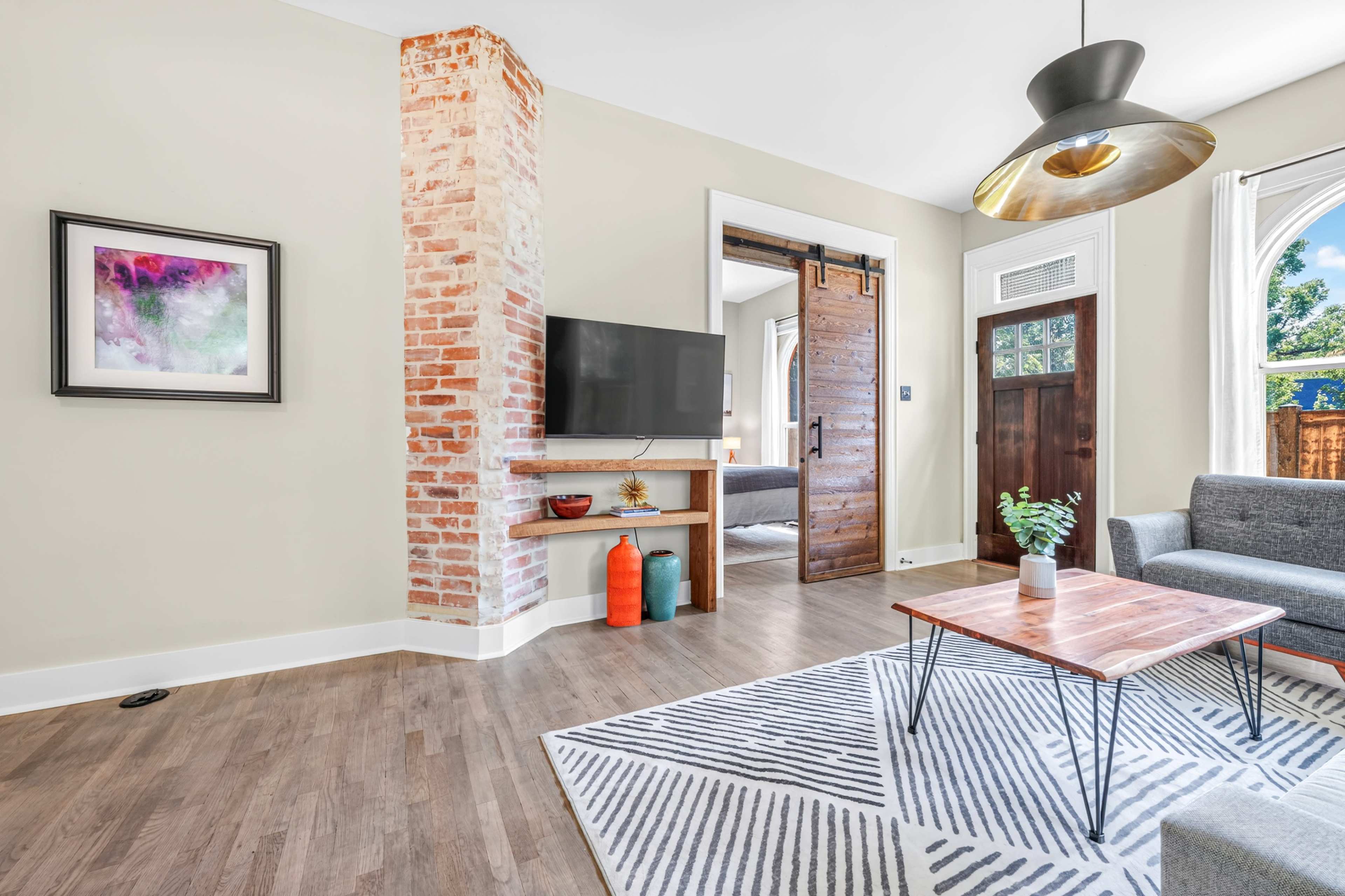The image shows a modern living room with a gray sofa, a wooden coffee table, a television mounted on a wall, and a front door leading to an outdoor area.