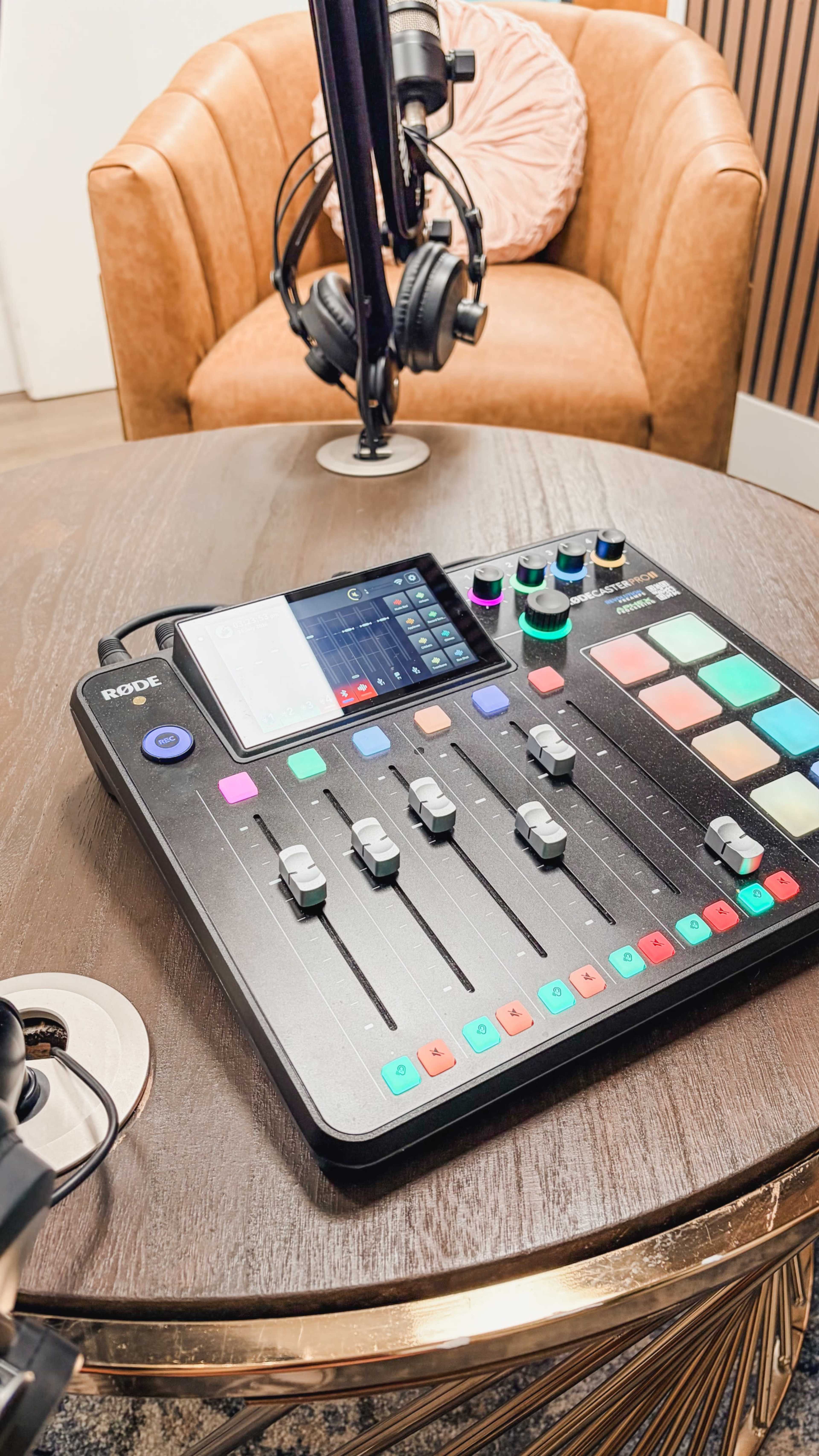 A digital audio mixer is placed on a wooden table, with headphones and a microphone visible in the background.