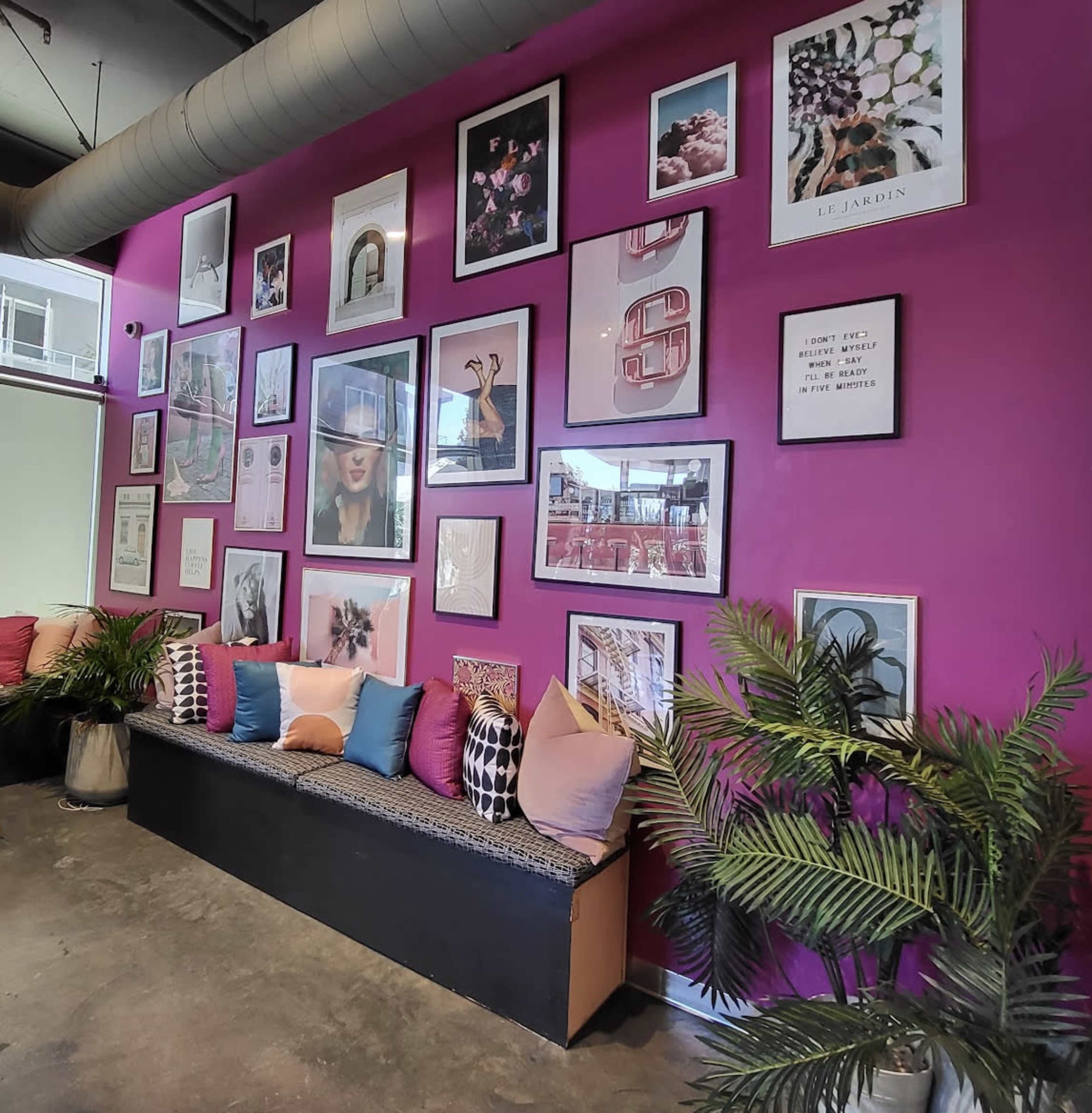The scene features a wall painted pink, adorned with various framed artworks and photographs, alongside a seating area with colorful cushions and potted plants.