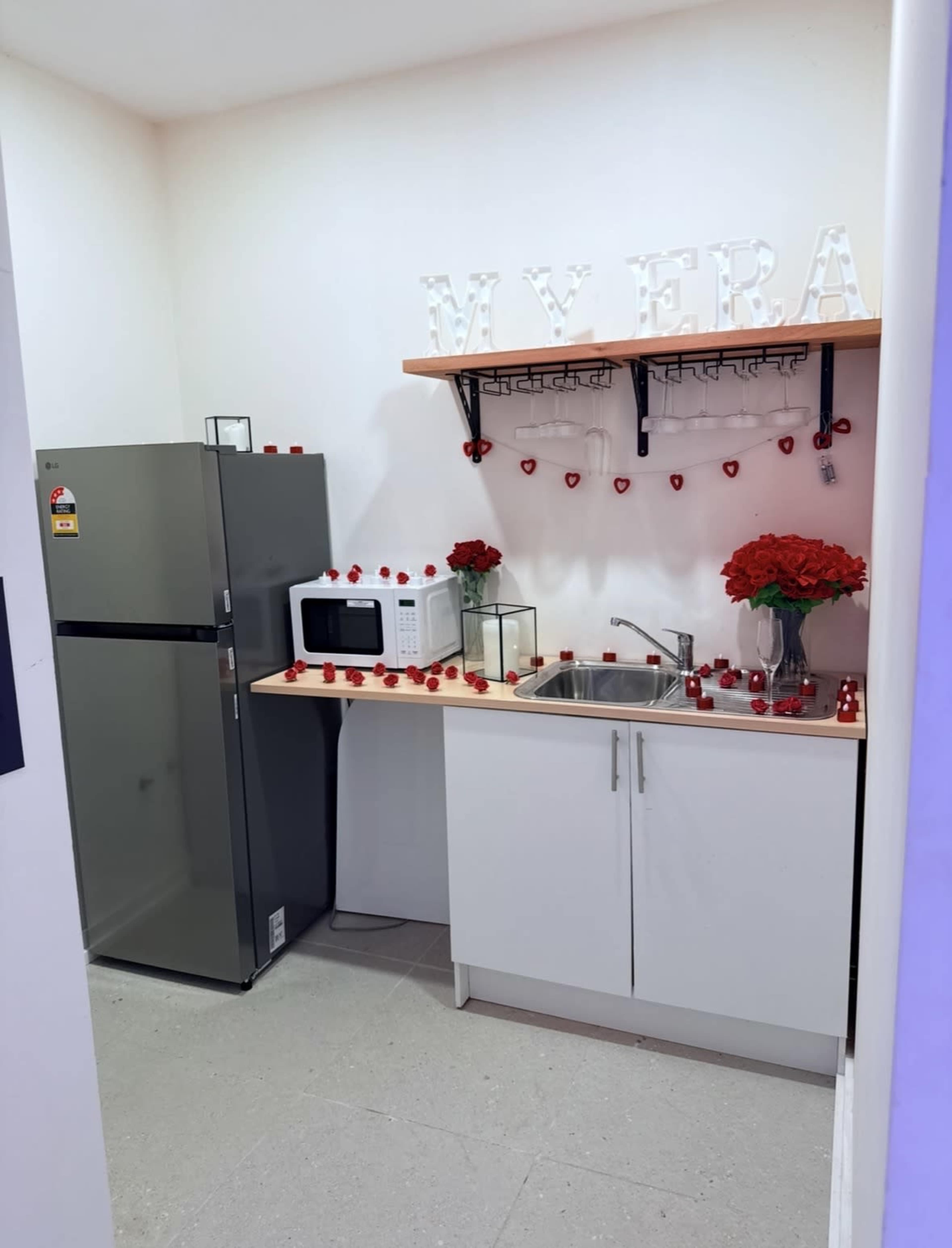 A small kitchen contains a refrigerator, microwave, and a sink, adorned with red decorations and flowers.