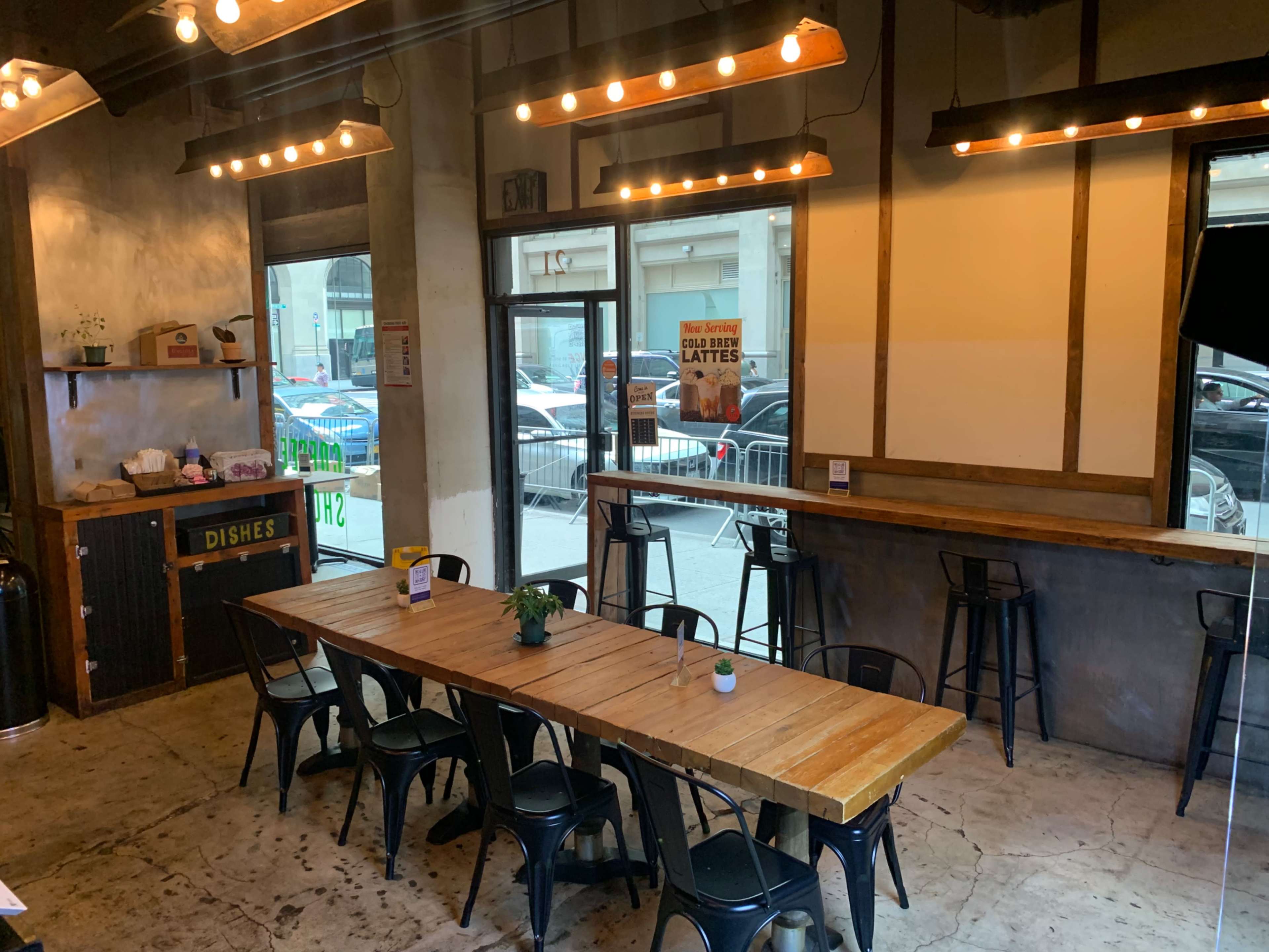 A spacious café interior featuring a long wooden dining table surrounded by black metal chairs, with large windows allowing natural light and a view of the street outside.