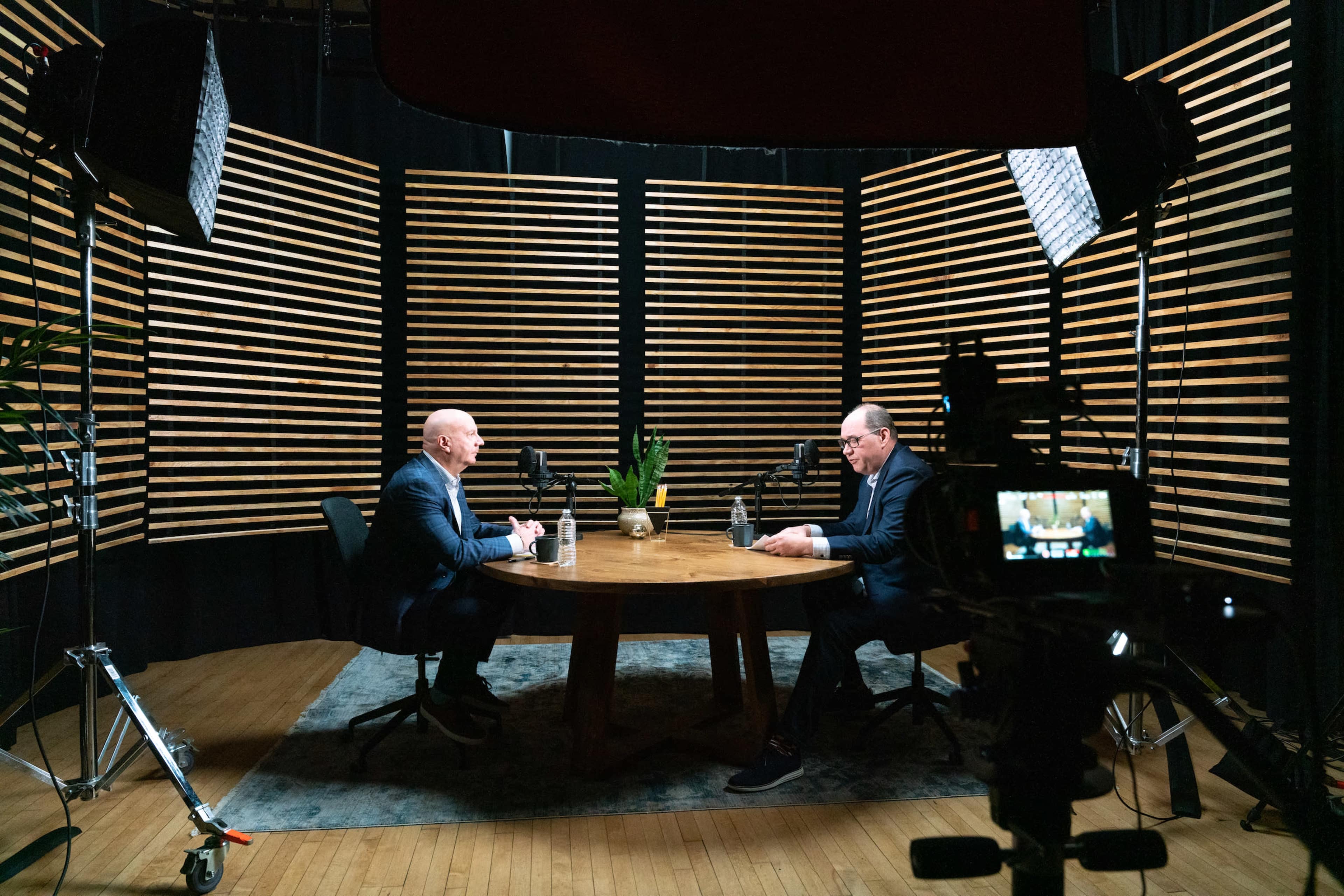 Two men are seated at a round wooden table in a studio with wooden slat walls, engaged in conversation, while cameras and lighting equipment are positioned nearby.