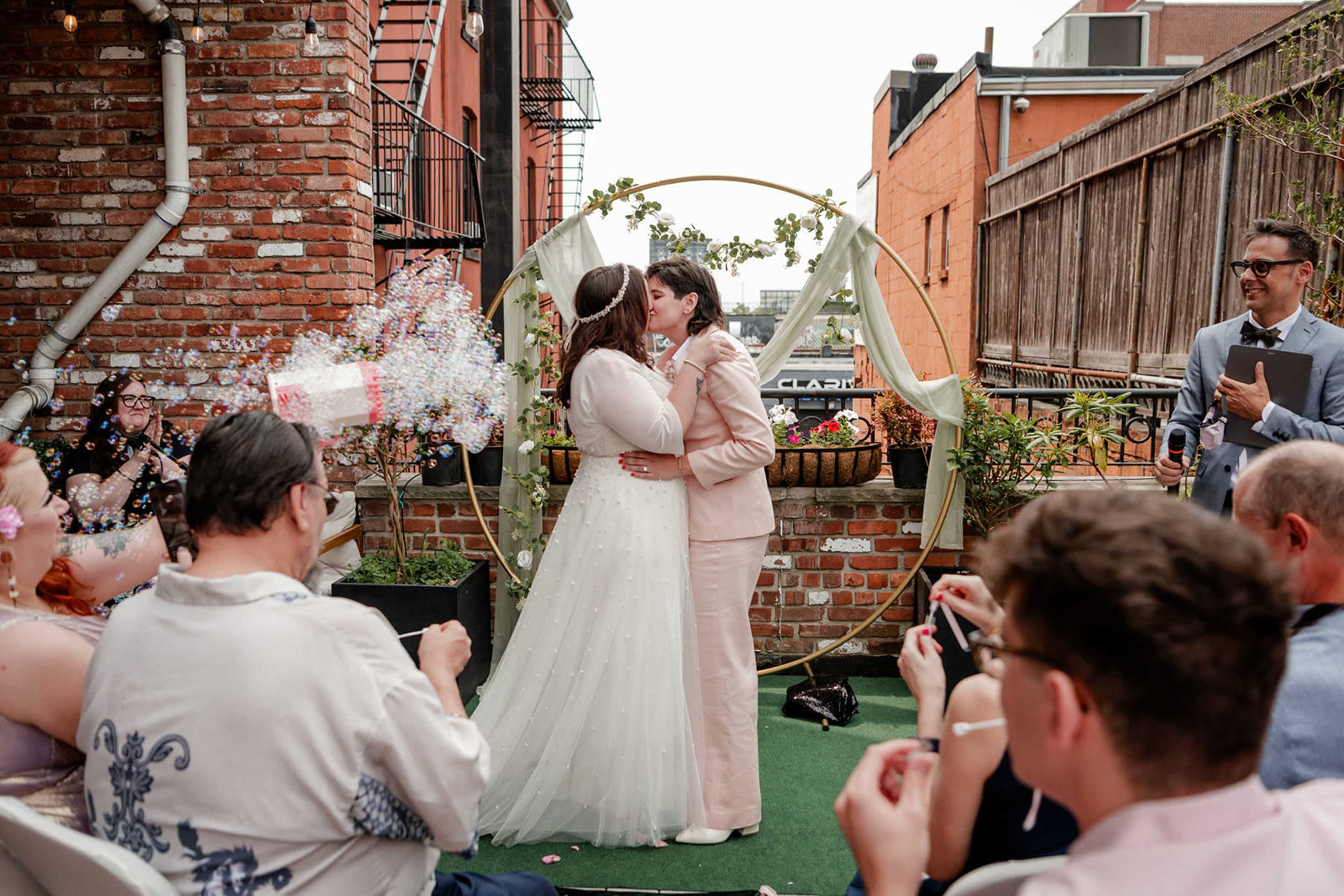 Two people in wedding attire kiss in an outdoor setting surrounded by guests, with a floral arch in the background.
