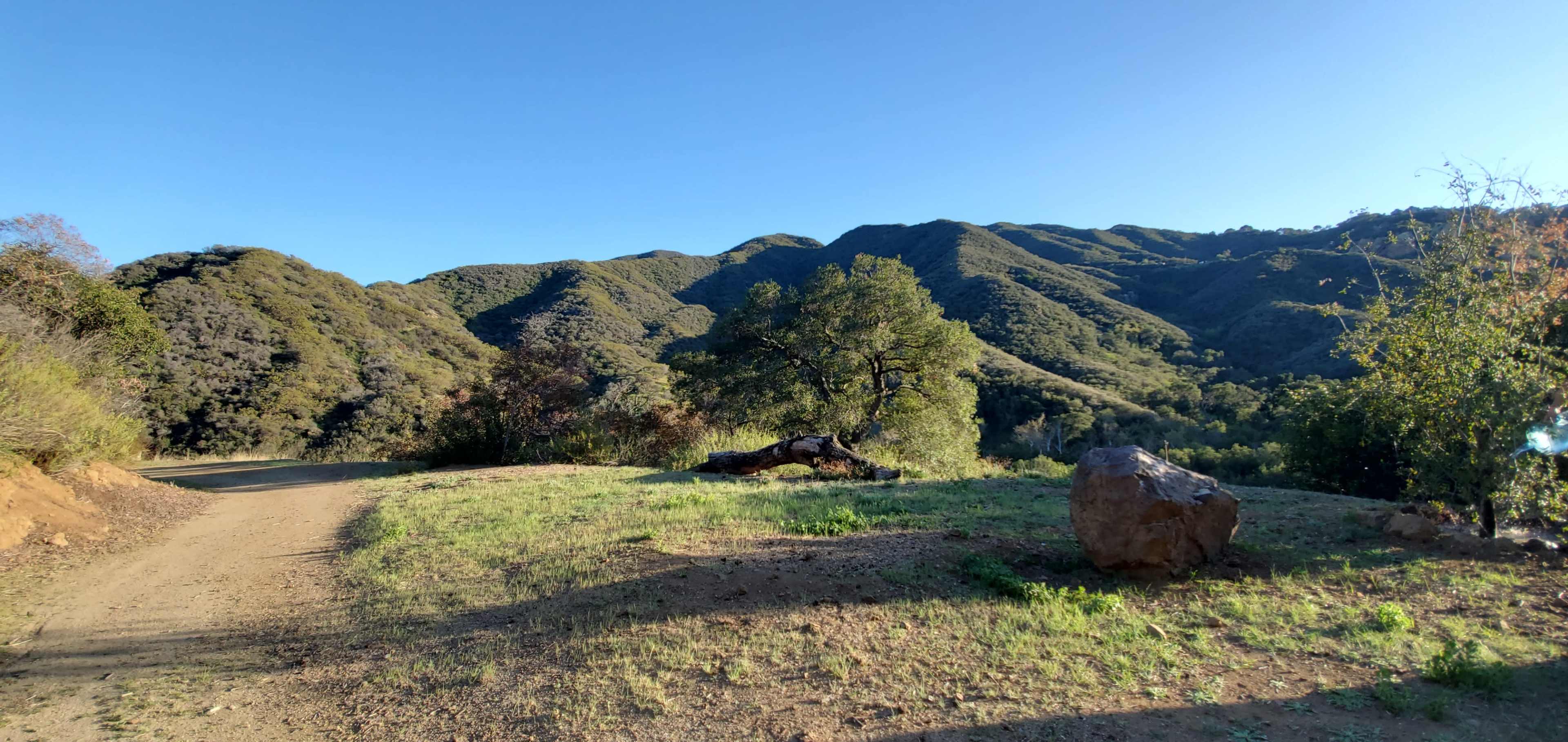 A winding dirt path leads through a grassy area surrounded by rolling green hills and a large rock in the foreground.