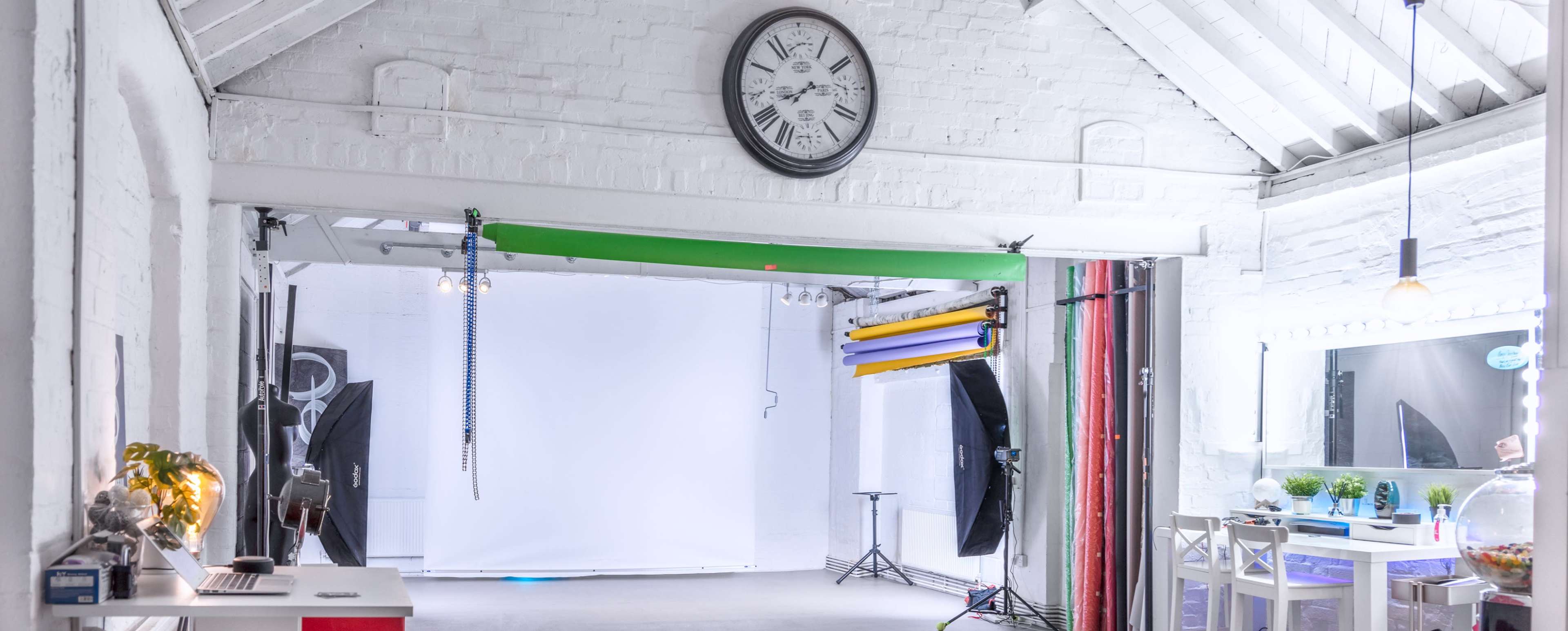 A brightly lit photography studio with a blank backdrop, lighting equipment, and a large clock on the wall.