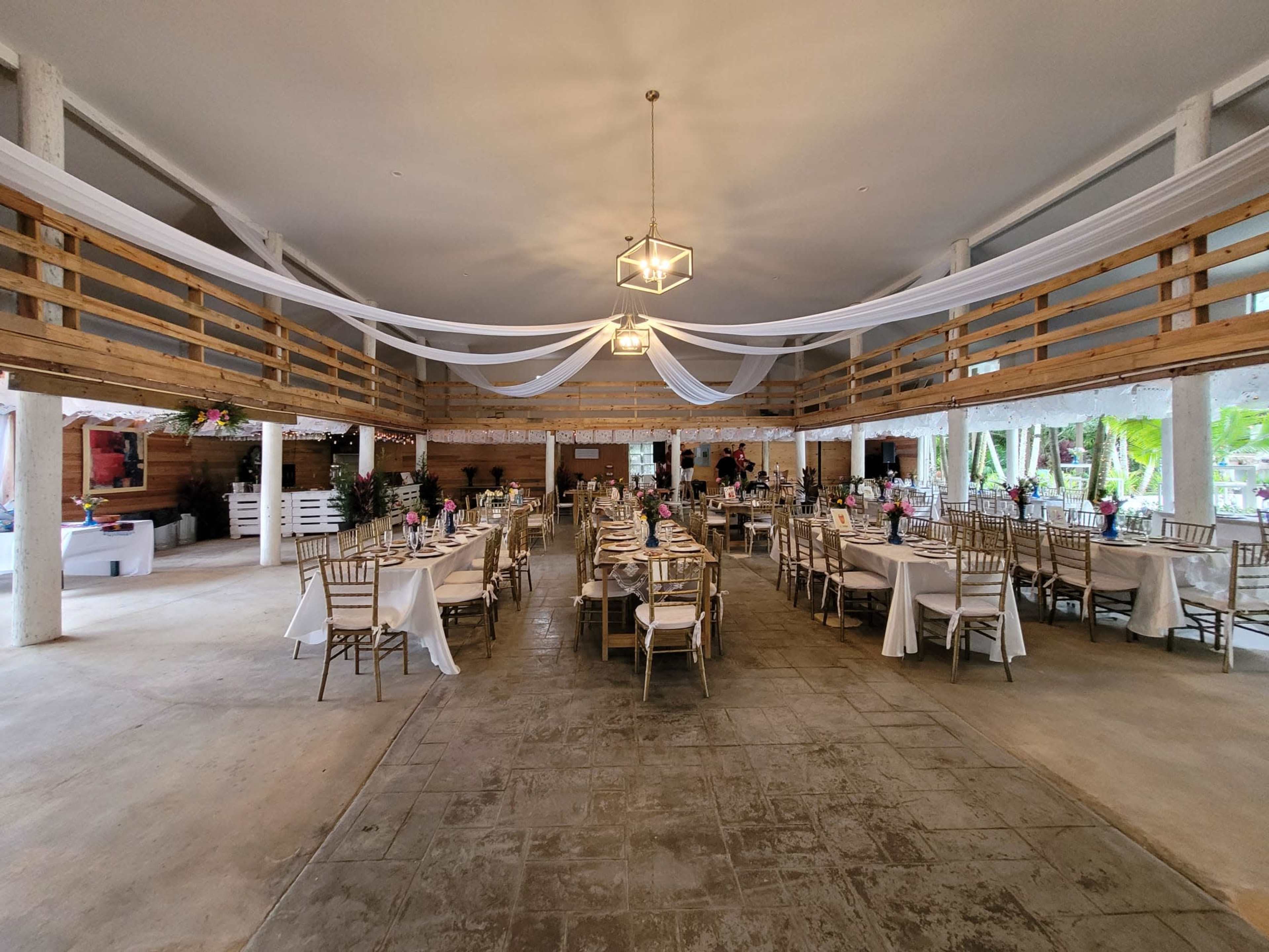 The image shows a spacious indoor event venue with neatly arranged tables covered in white linens and decorations, featuring a rustic wooden decor and a high ceiling with draped fabric.