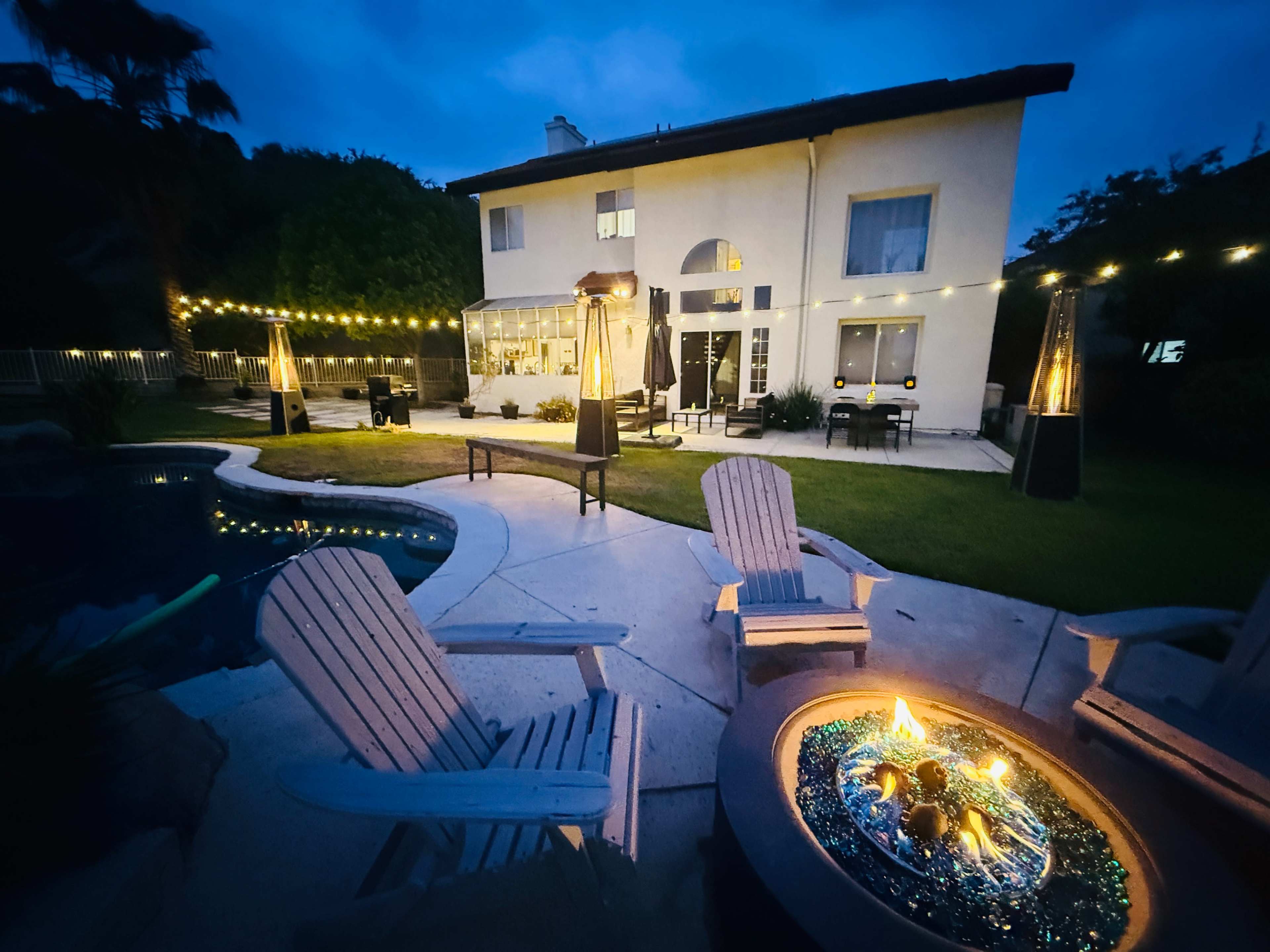 The scene depicts a backyard at night featuring a swimming pool, a fire pit with flames, and a well-lit house surrounded by string lights and patio furniture.