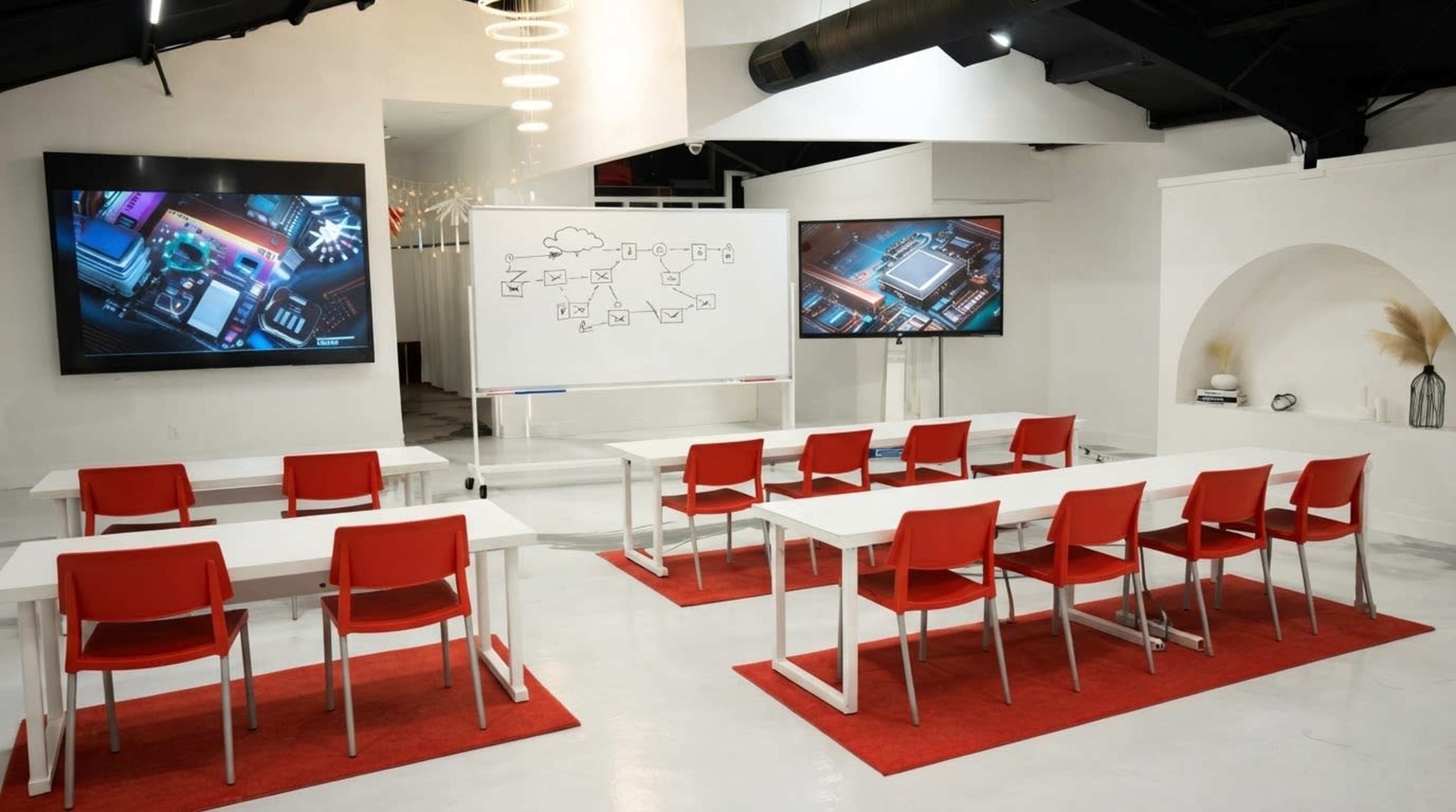 A modern conference room features two large screens displaying digital visuals, a whiteboard with diagrams, and rows of red chairs arranged around white tables.
