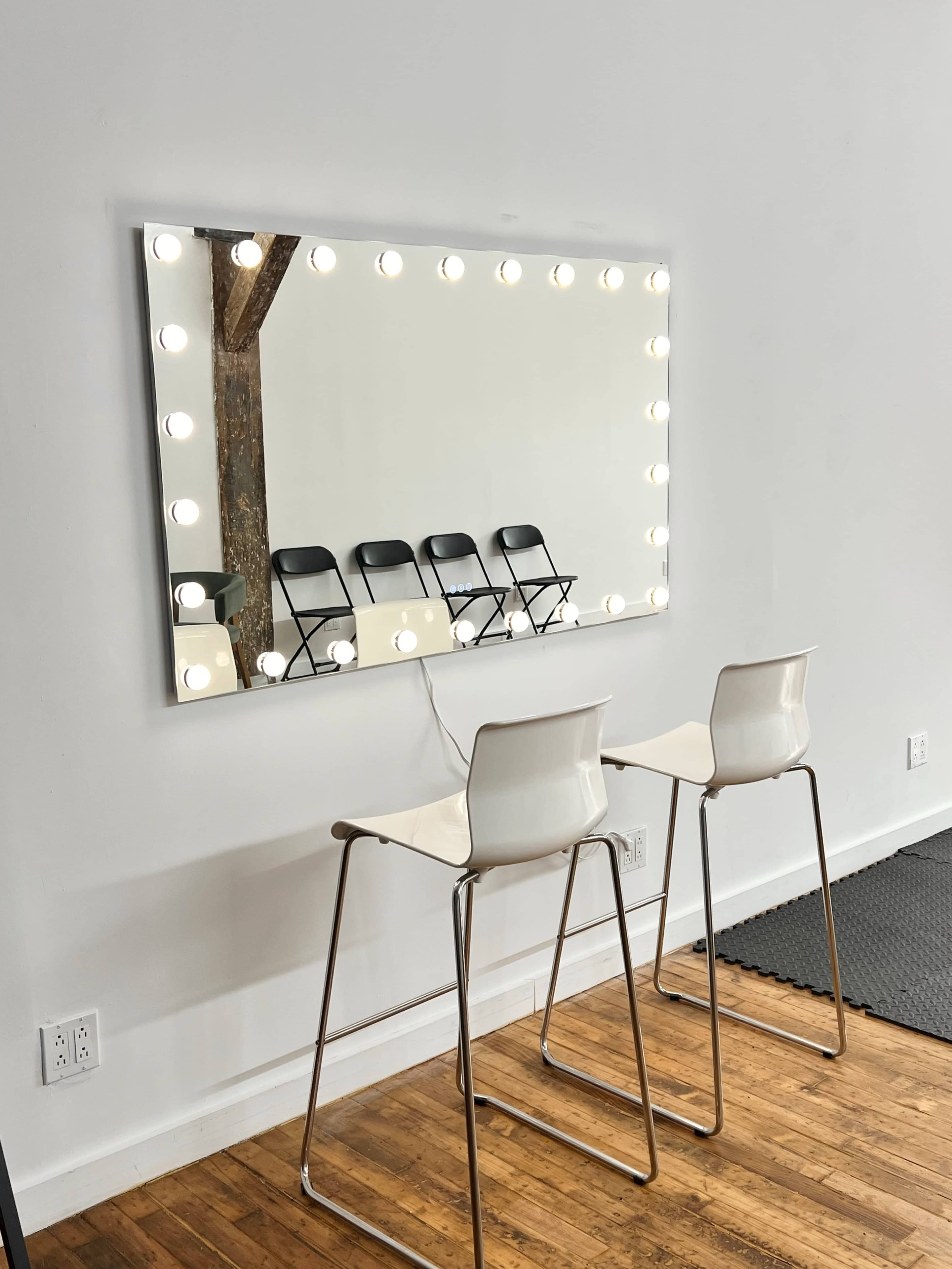 A large illuminated mirror hangs on a wall above two white bar stools, with several black chairs neatly arranged in the background.