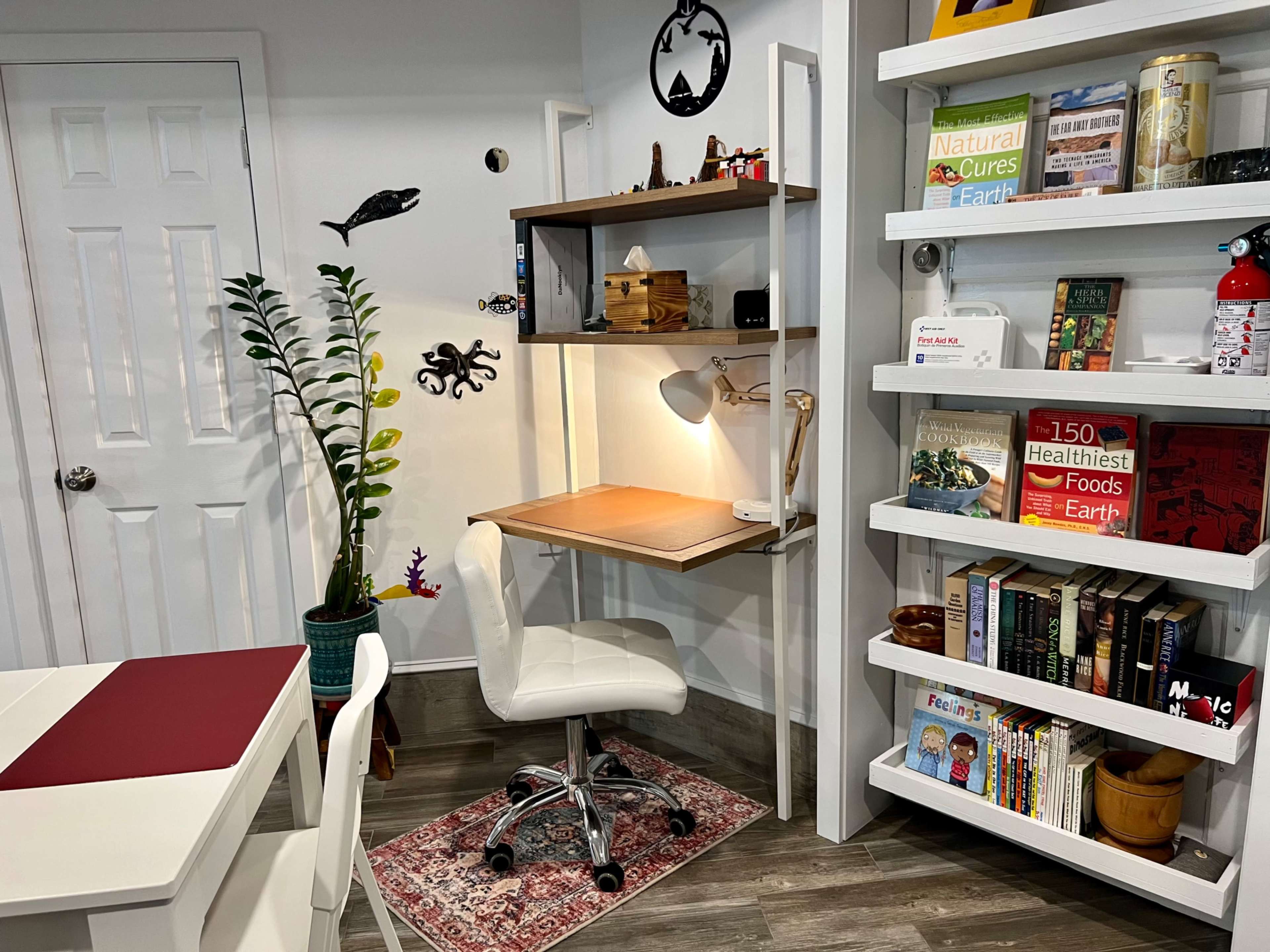 The image shows a compact workspace with a wooden desk, a swivel chair, a plant, and shelves filled with books and decorative items.