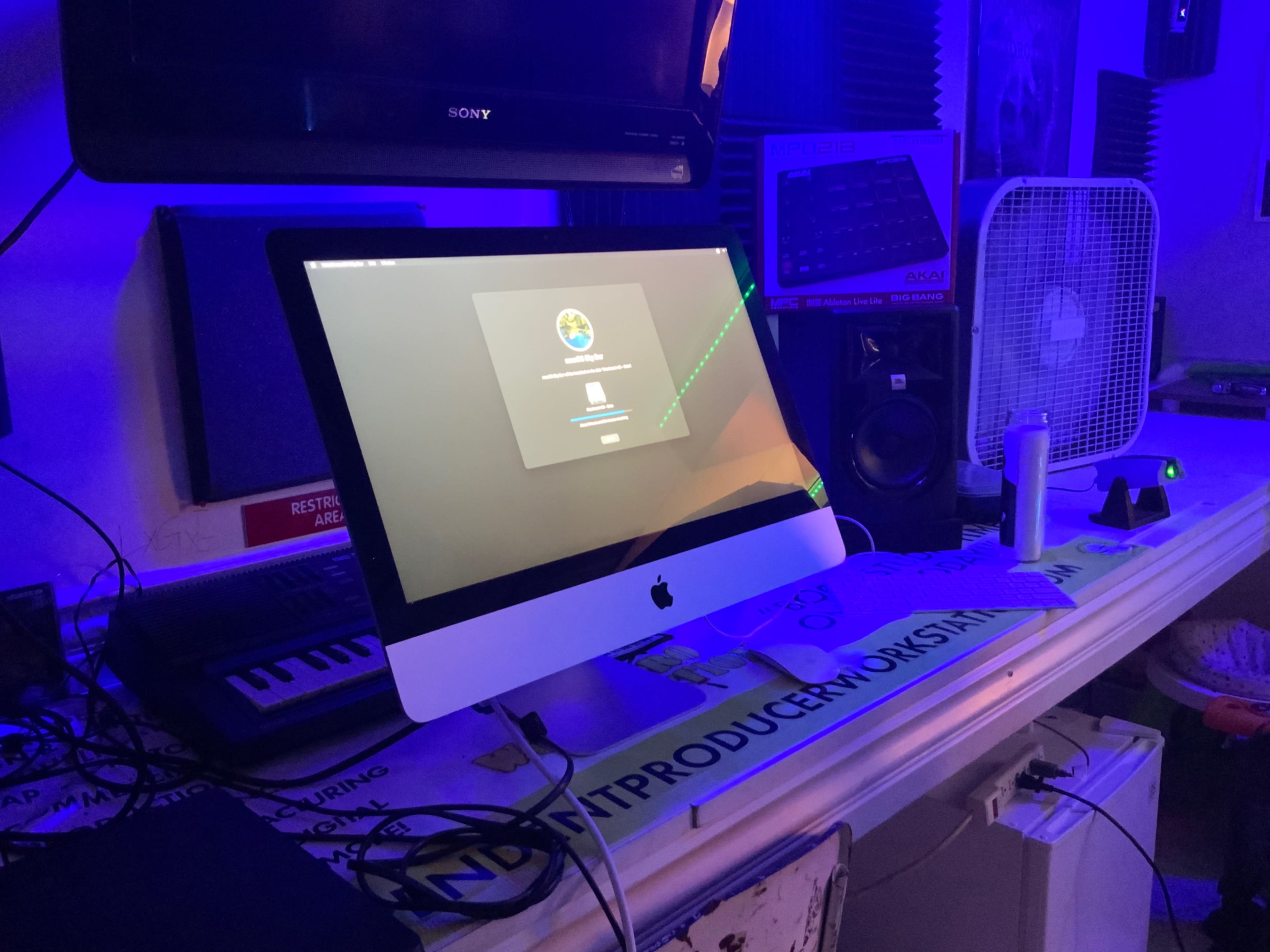 A lit-up iMac sits on a cluttered desk surrounded by music equipment and studio decor.