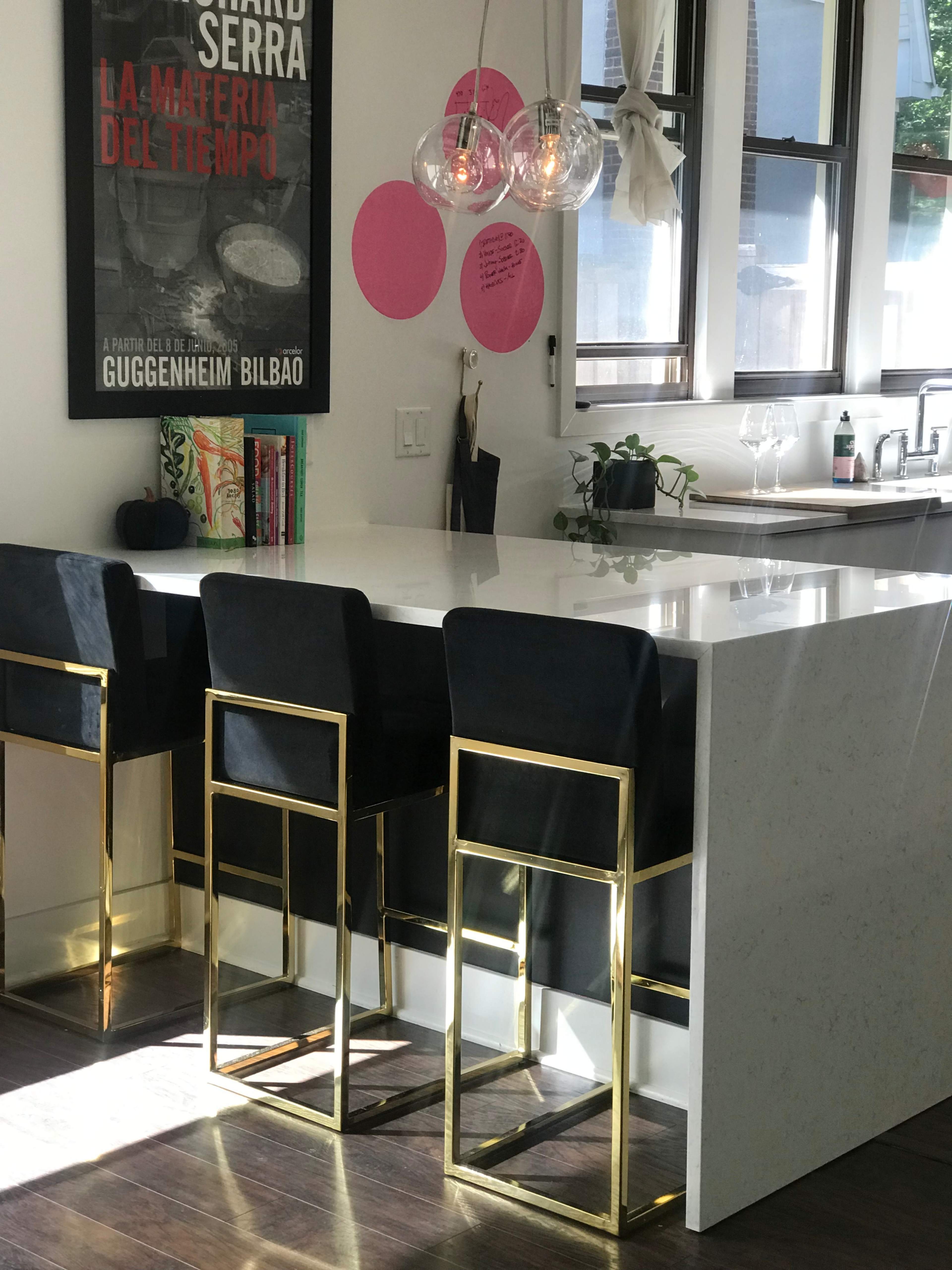 A sleek kitchen island with four black bar stools featuring gold frames sits under modern pendant lights in a well-lit space adorned with artwork.