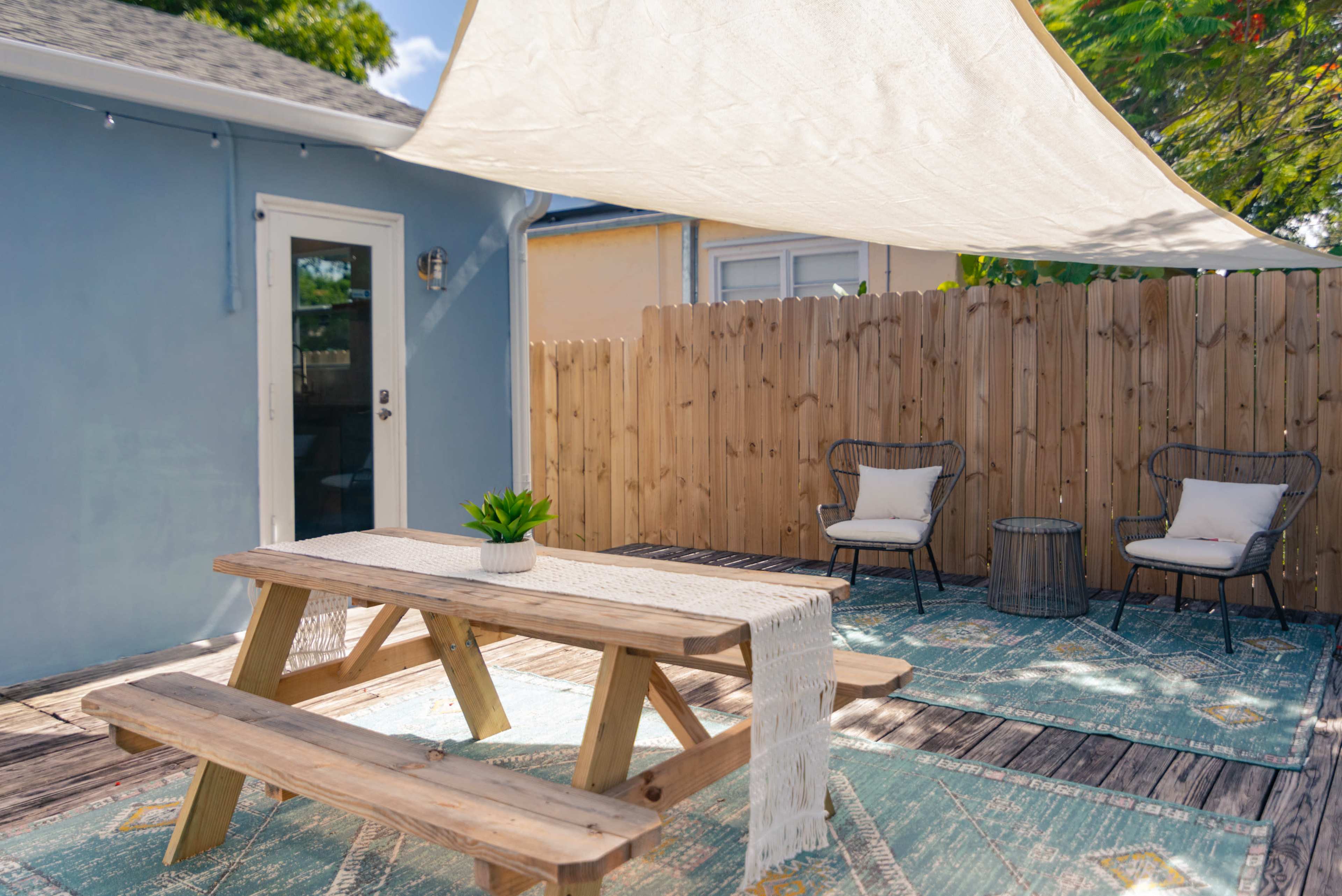 A wooden picnic table under a shaded area with two chairs and a small table on a wooden deck surrounded by a fenced yard.