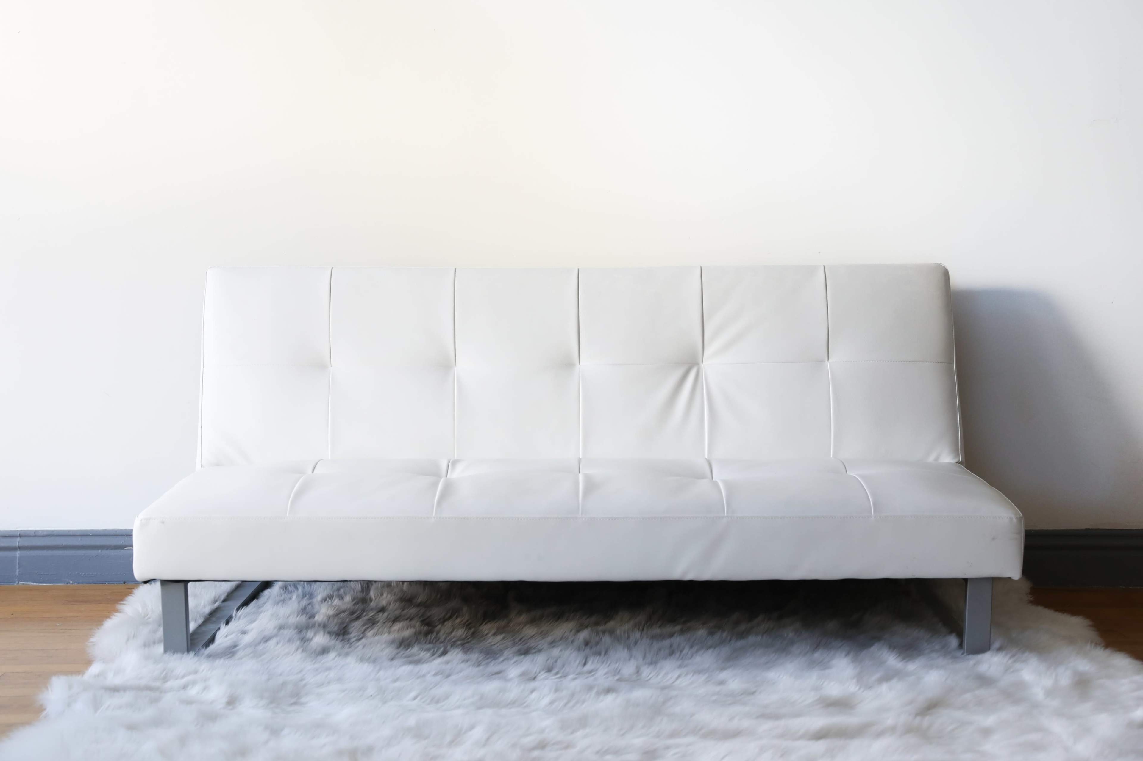 A white futon is positioned on a fluffy rug against a plain wall.