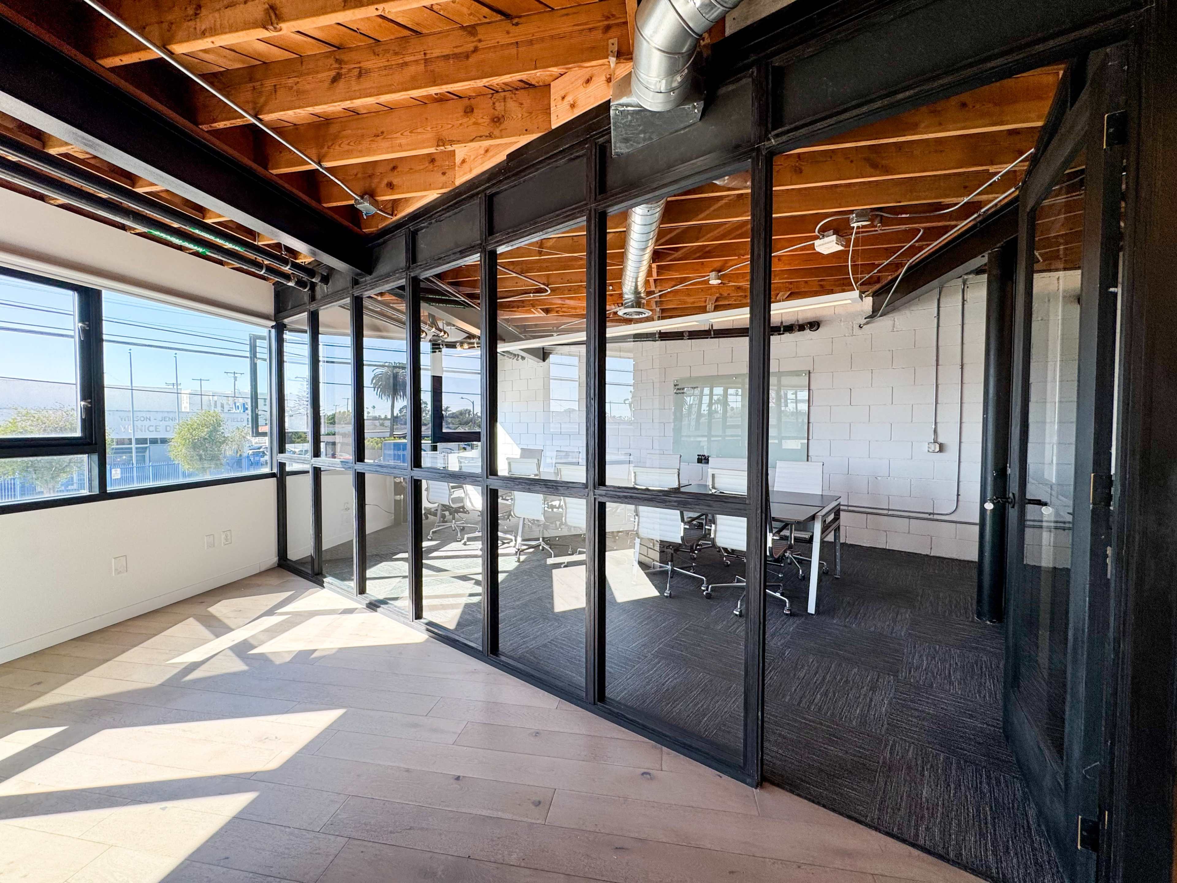 The image shows a modern office space featuring a glass-walled conference room with a table and chairs, set against a backdrop of exposed wooden beams and a light-colored floor.
