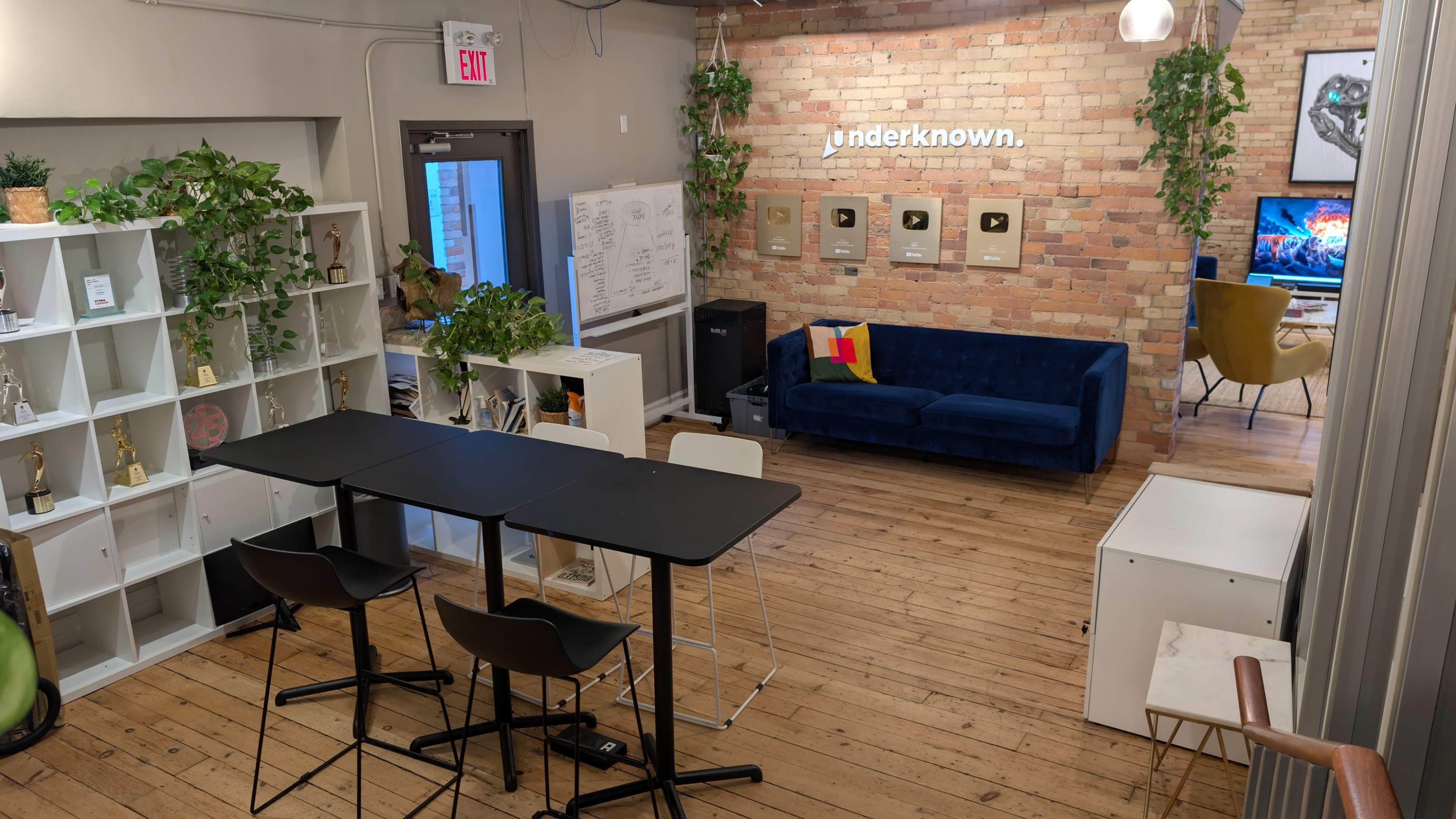 The image shows a modern workspace featuring a seating area with a blue couch, a dining table with chairs, and an exposed brick wall displaying framed plaques.