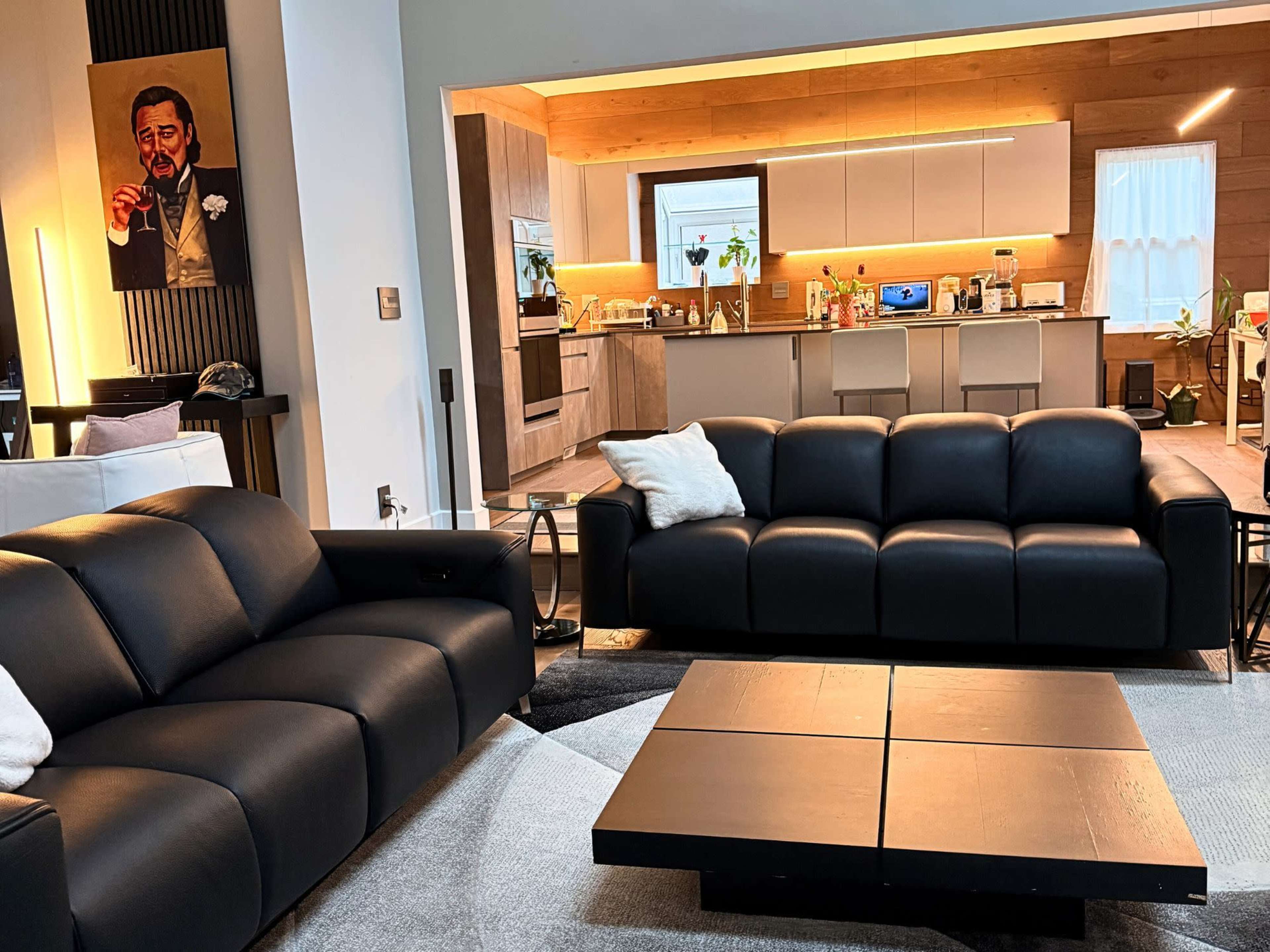 The image shows a modern living room featuring black leather sofas and a large coffee table, with a kitchen area visible in the background.