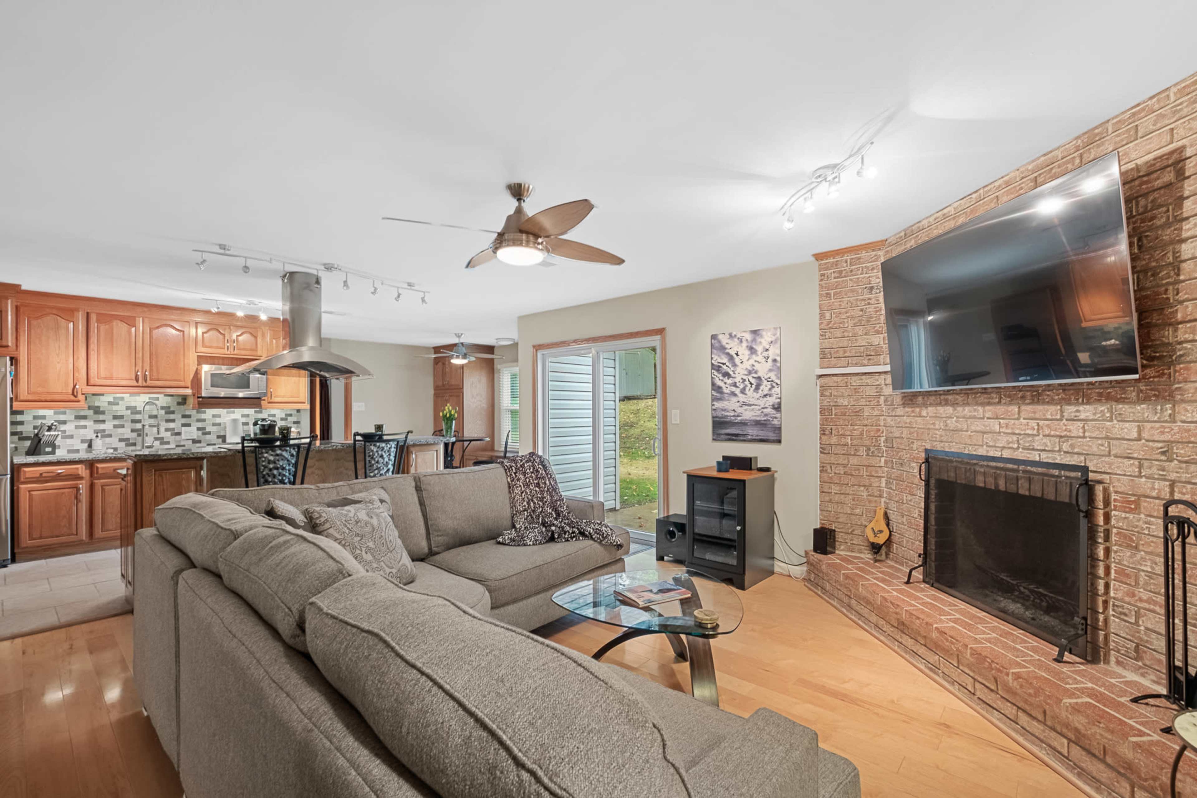 A living room with a grey sectional sofa, a brick fireplace, and a television mounted on the wall, adjacent to an open kitchen area with wooden cabinetry.