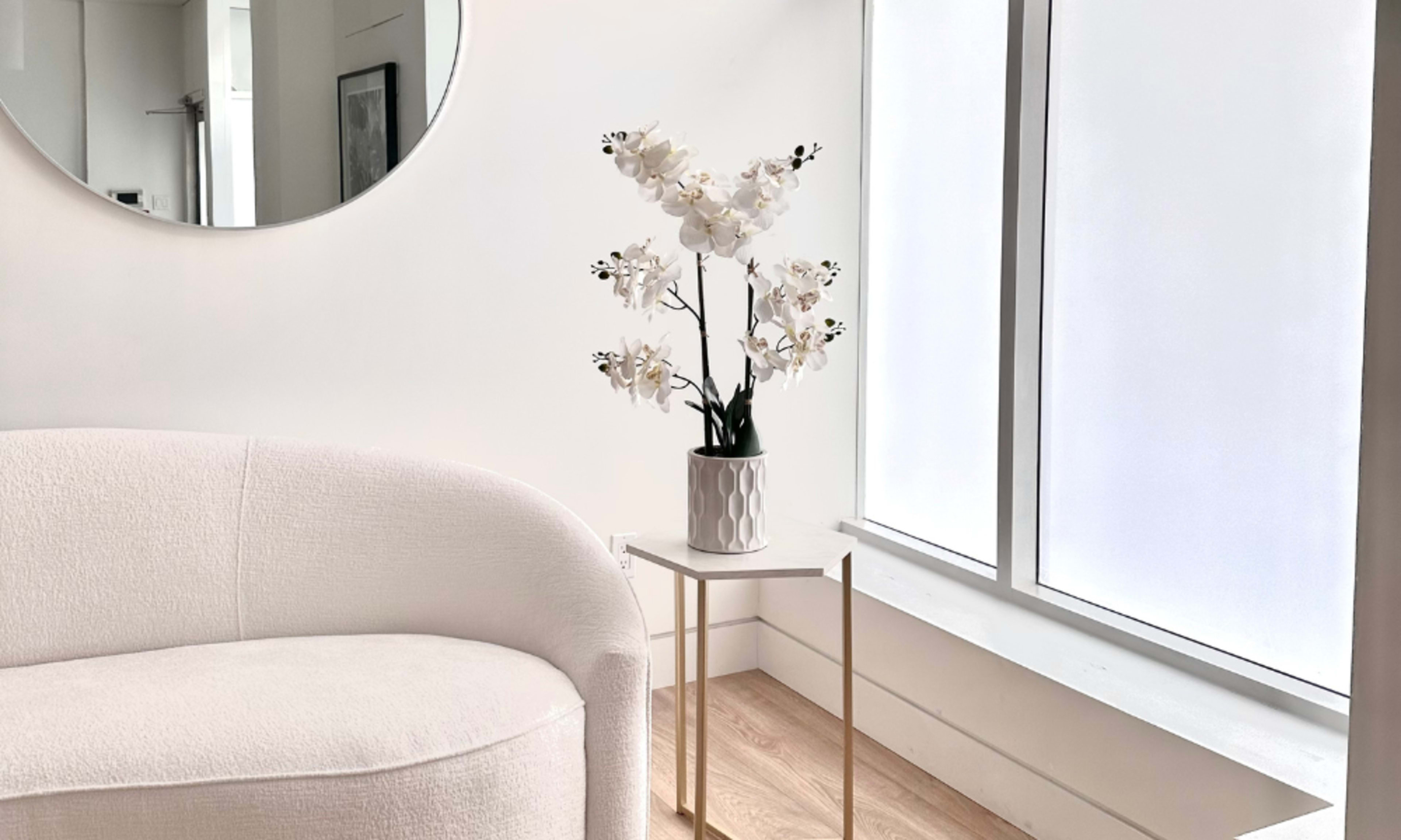 A white sofa sits next to a flowering orchid arrangement on a small table beside a large window.