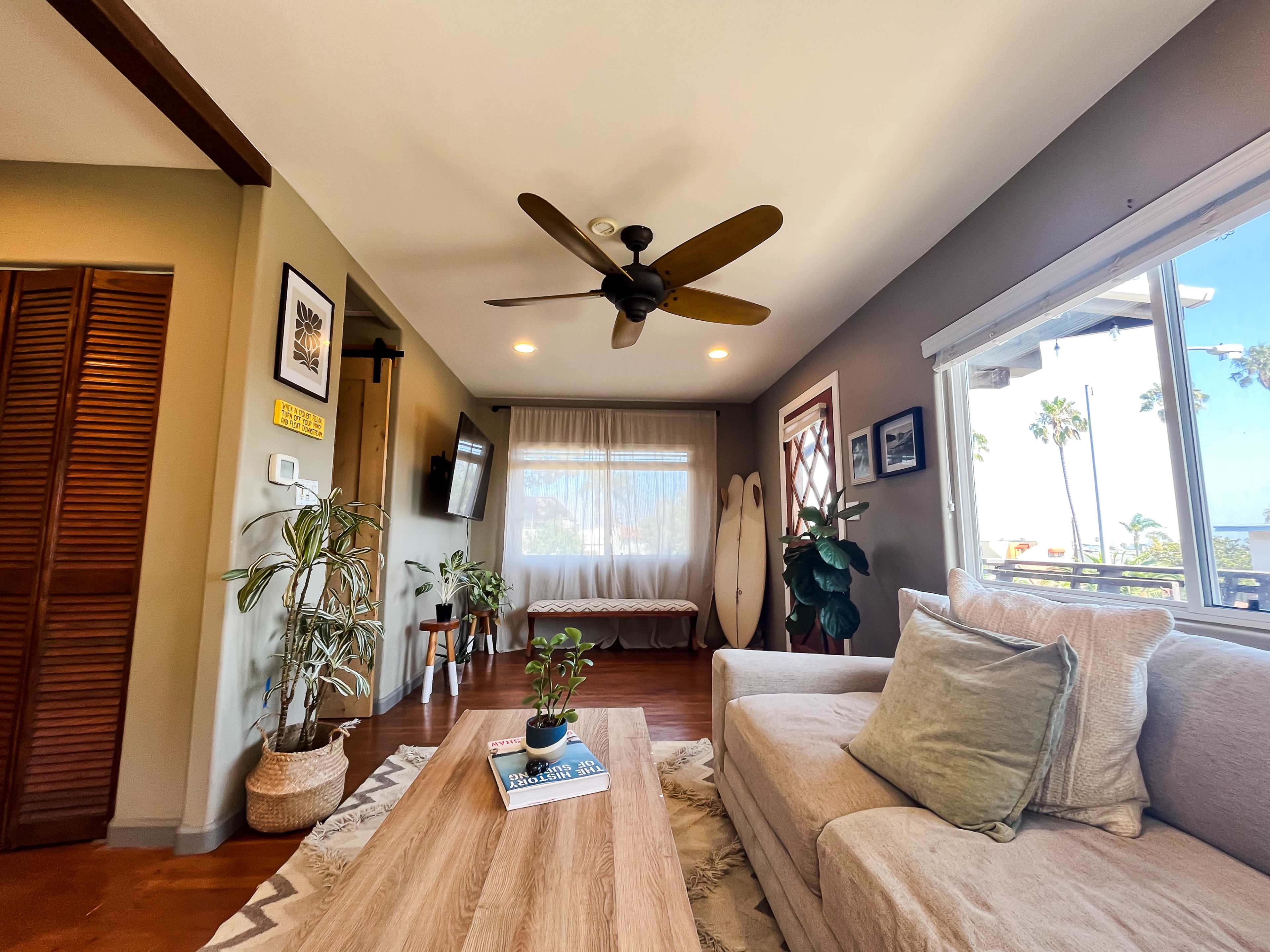 A cozy living room features a beige sofa, wooden coffee table, plants, a ceiling fan, and large windows with a view of palm trees.