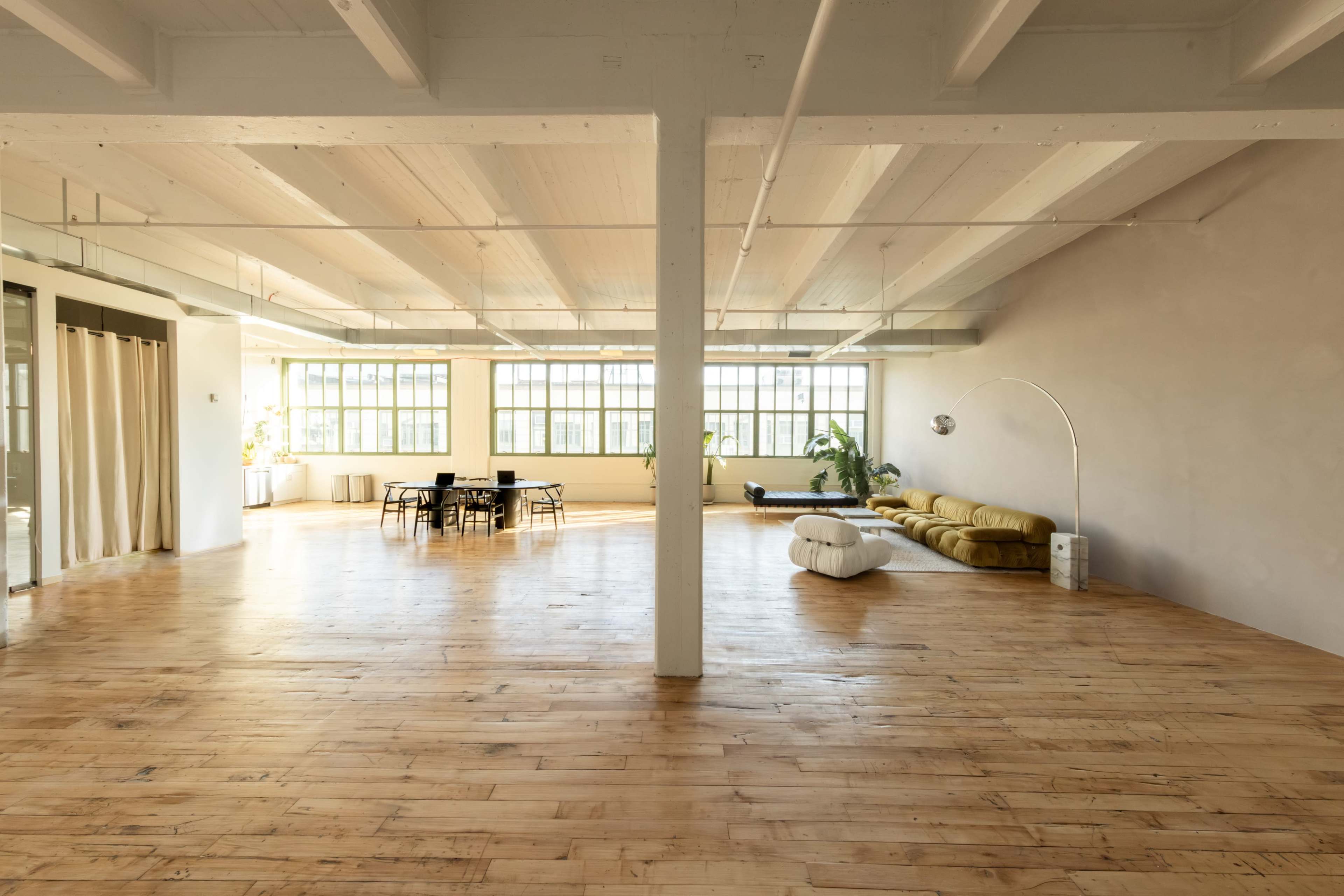 The image shows a spacious, well-lit interior featuring wooden floors, large windows, a yellow sofa, a round floor lamp, and a dining table with chairs.