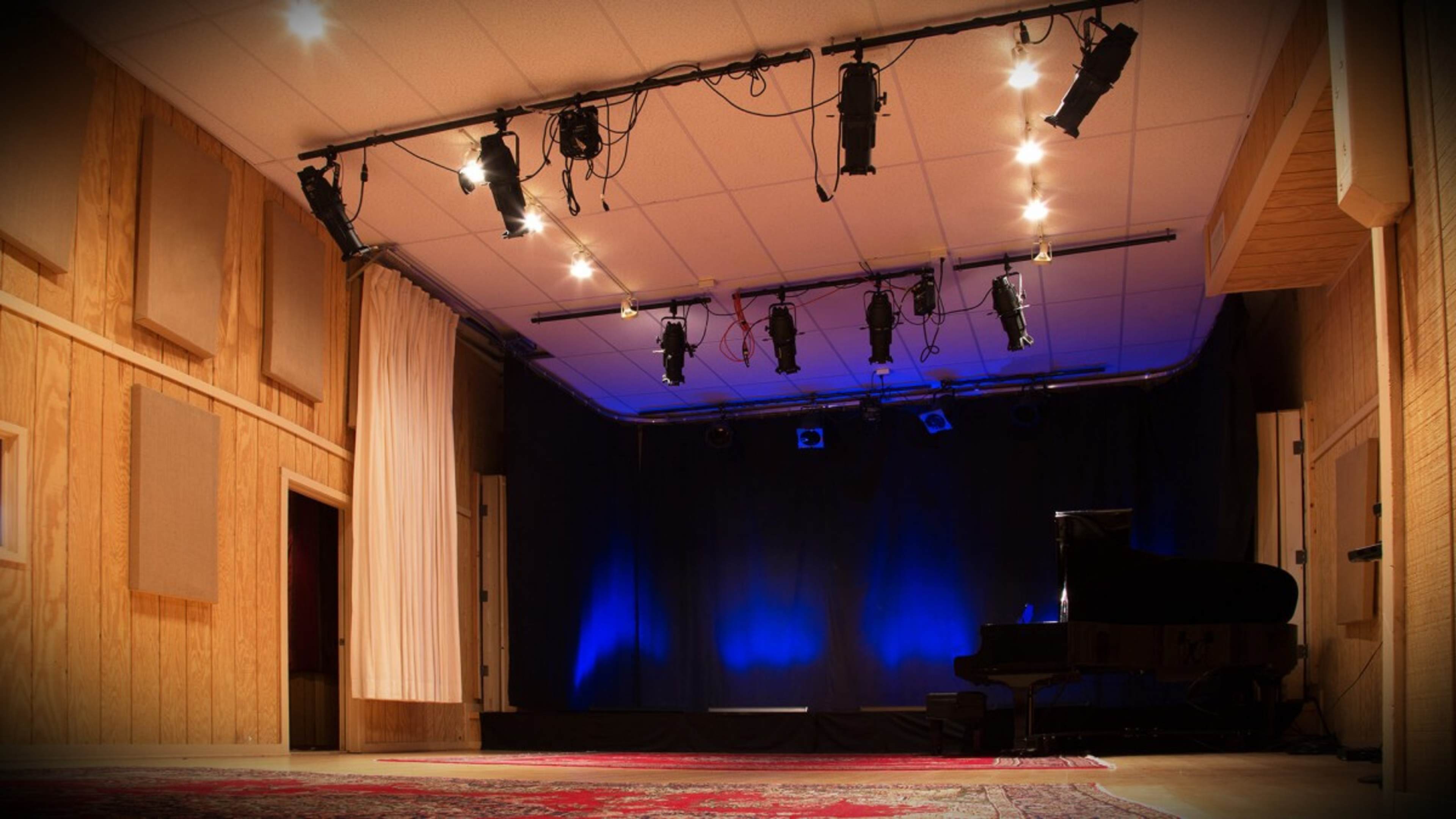 A sparsely decorated stage with a grand piano and blue lighting, set in a room with wooden paneling and soundproofing panels.