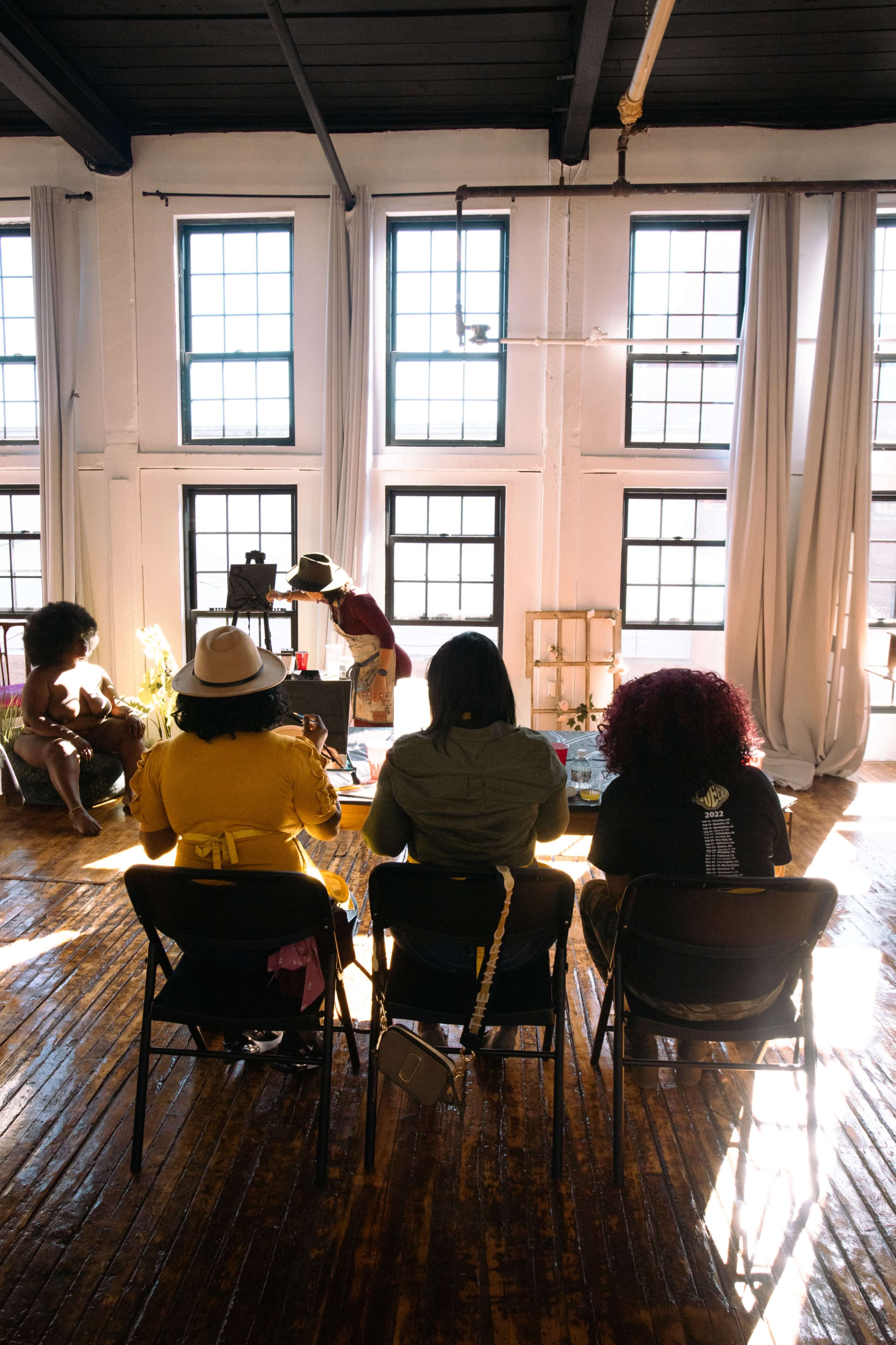 Four individuals sit in chairs facing a woman working at a table in a well-lit room with large windows and wooden floors.