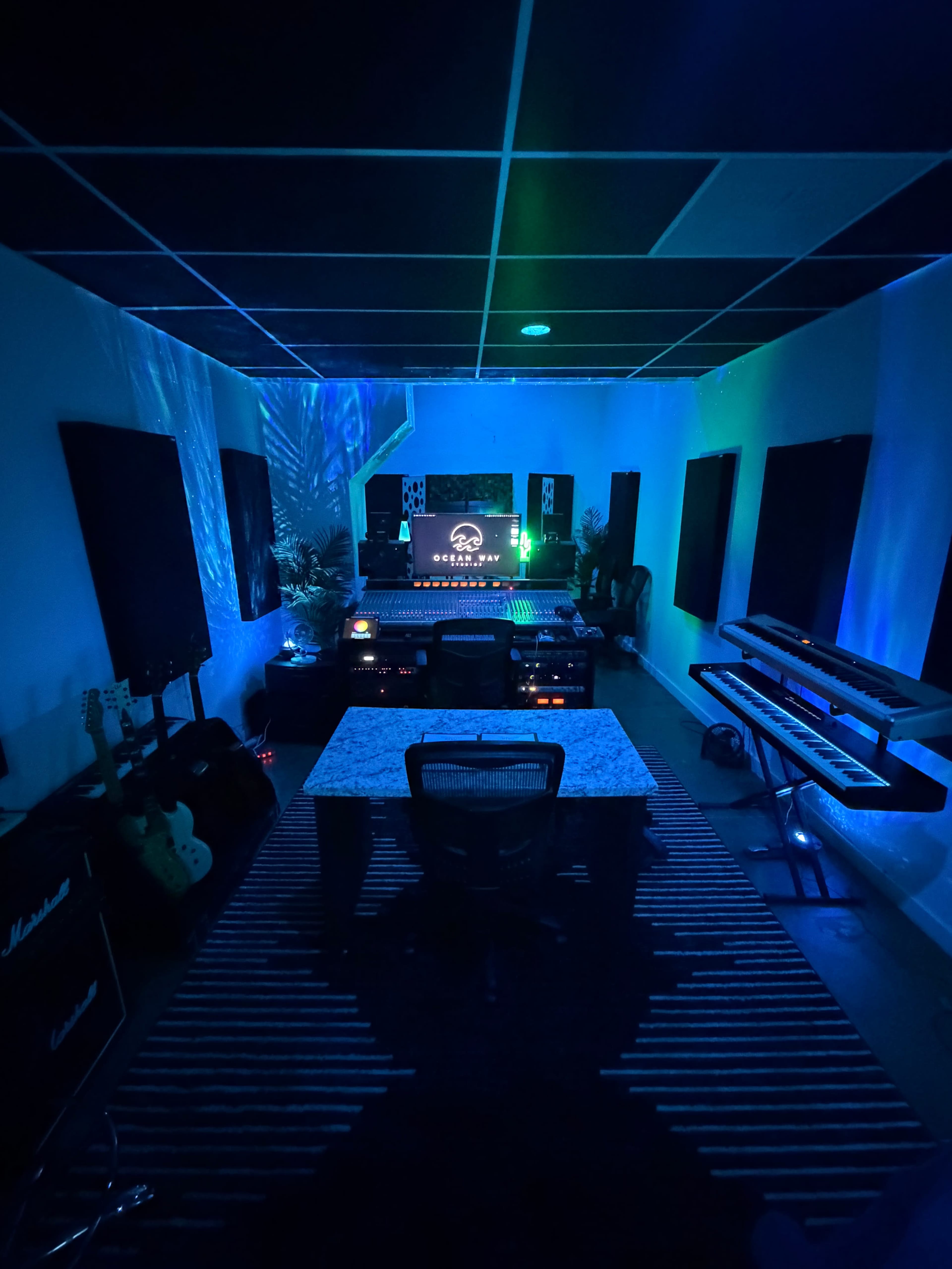 The image shows a dimly lit music studio featuring a central desk with recording equipment, guitars, and a keyboard along the walls, illuminated by blue and green lights.